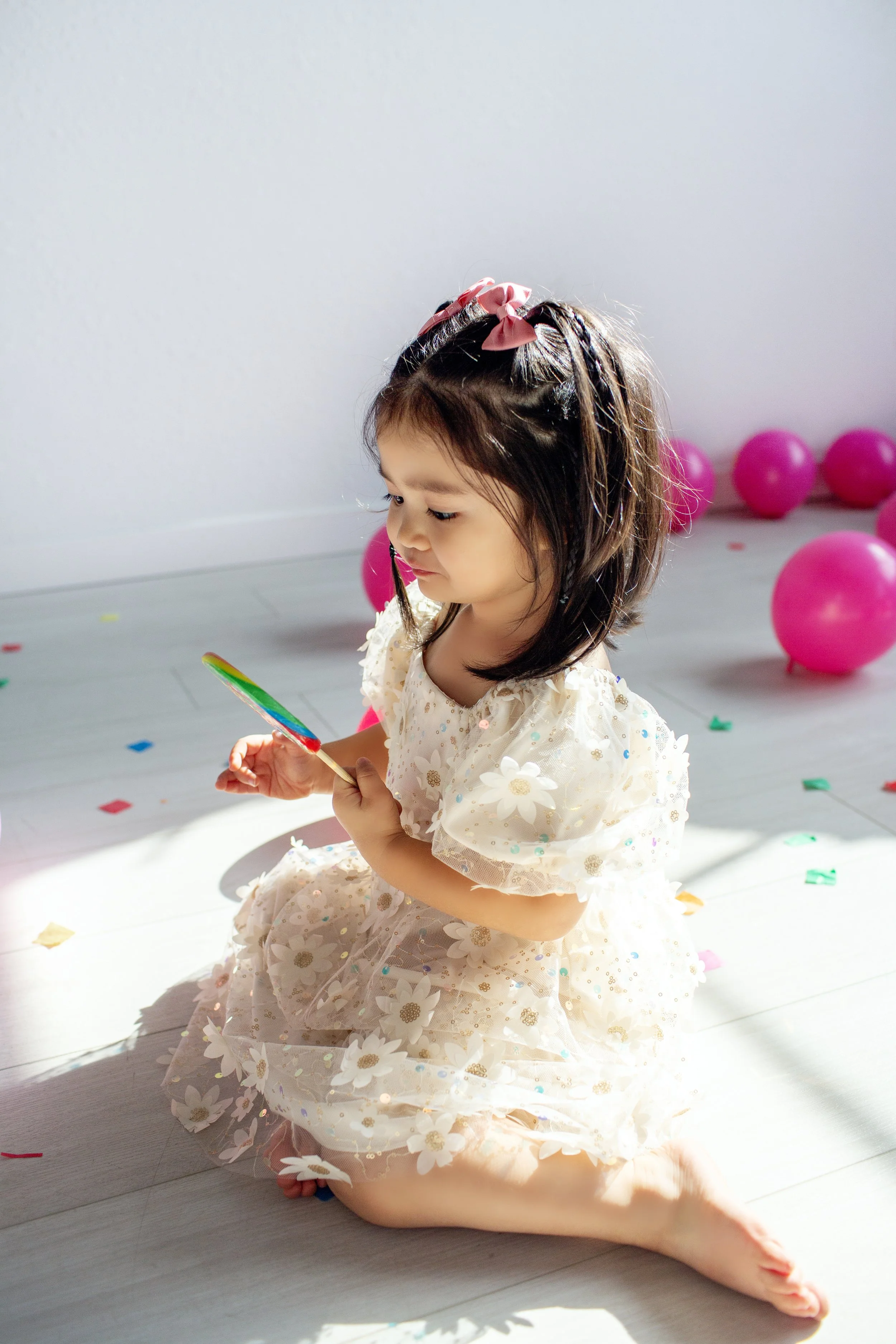 A young girl kneeling on the floor holding a rainbow-colored fan at a celebration with pink balloons and scattered confetti.
