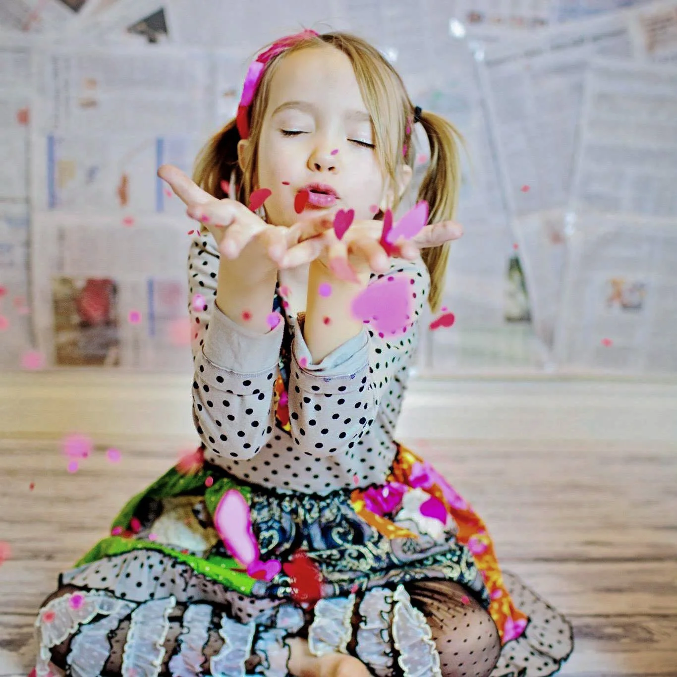 A young girl with pigtails blowing pink heart-shaped confetti in the air, seated on the floor with a background of newspapers taped to the wall.