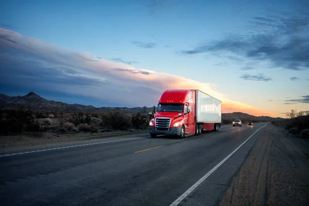Red semi-truck driving on a desert highway at sunset with distant mountains.