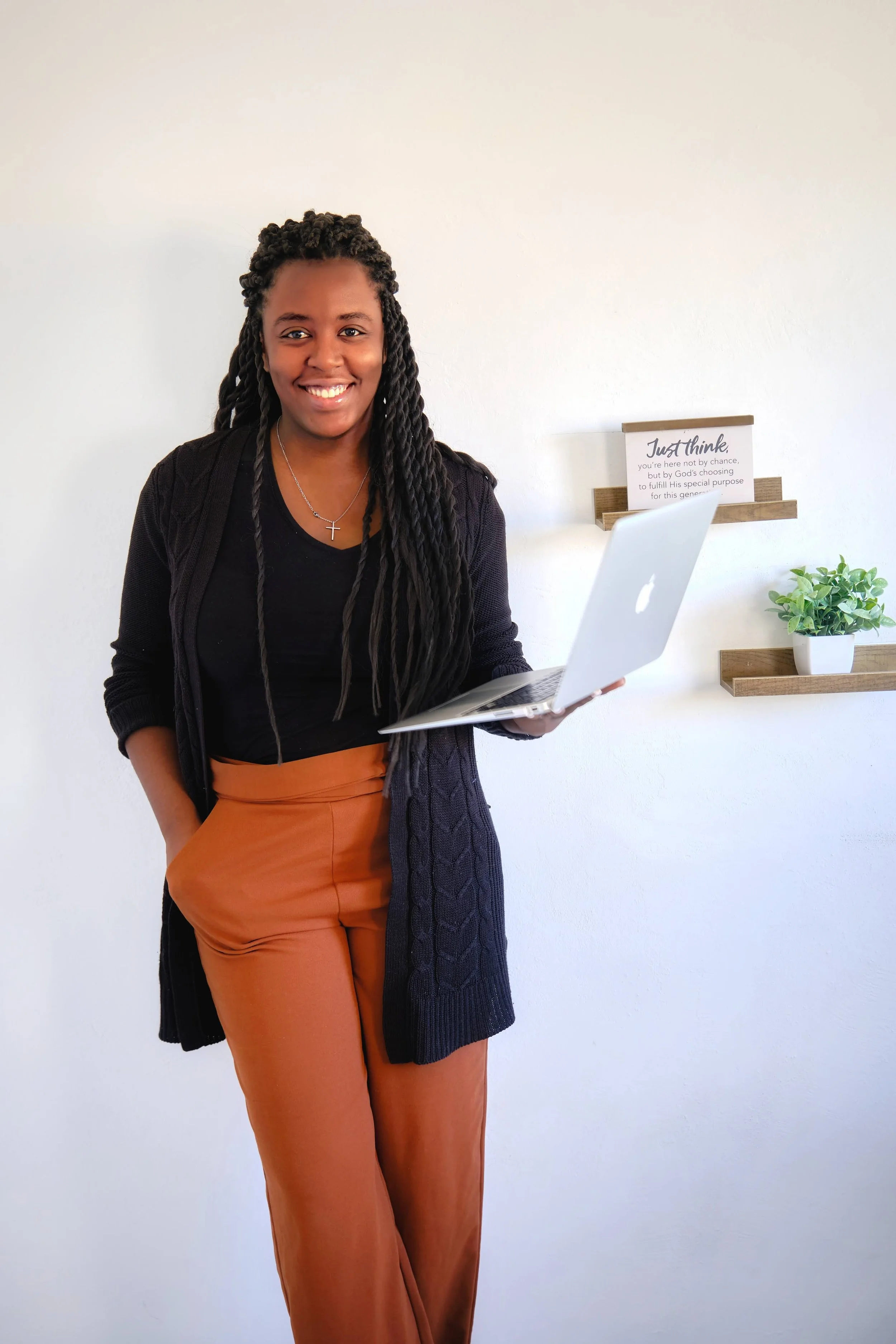 A woman with long braided hair smiling while holding an open laptop, standing indoors against a white wall with decorative shelves and a plant.