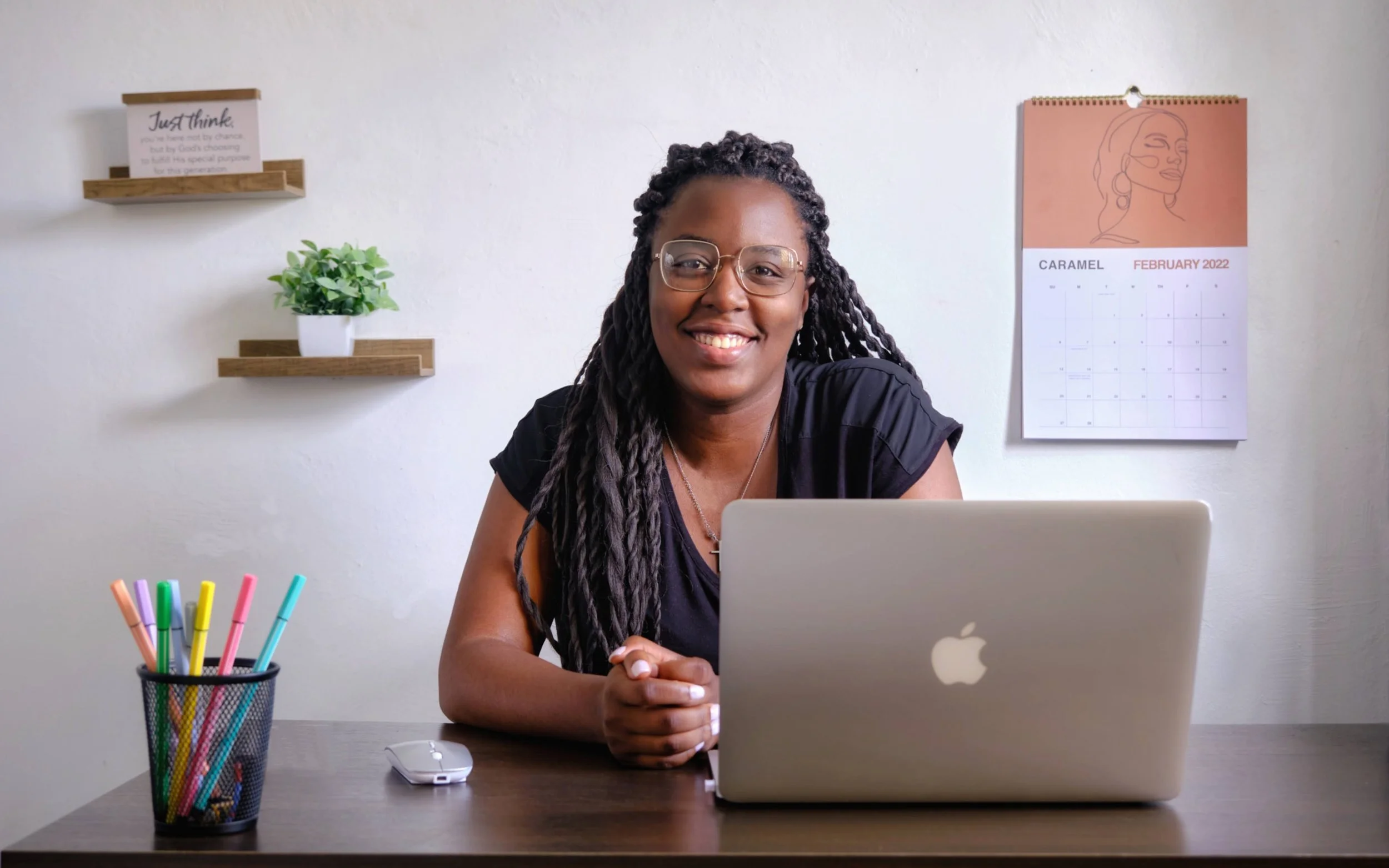A smiling woman with glasses and long braided hair sitting at a desk with a silver MacBook, a wired mouse, and a pencil holder with colorful pens. The background is a white wall with two wooden shelves, a small green plant, and a wall calendar for February 2022.