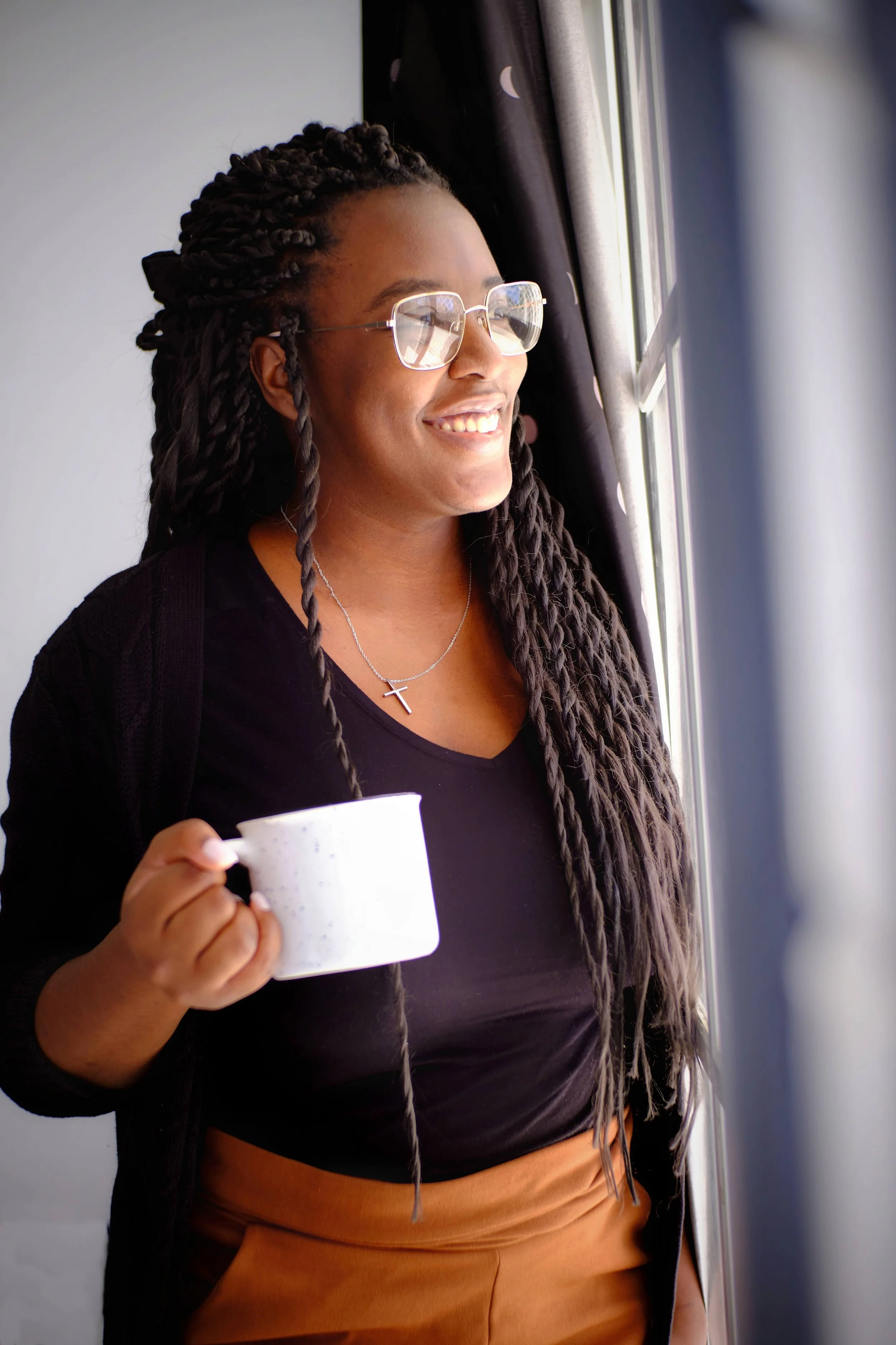 Bisi with long, twisted hair and glasses smiling while holding a white speckled mug, standing by a window with black curtains.