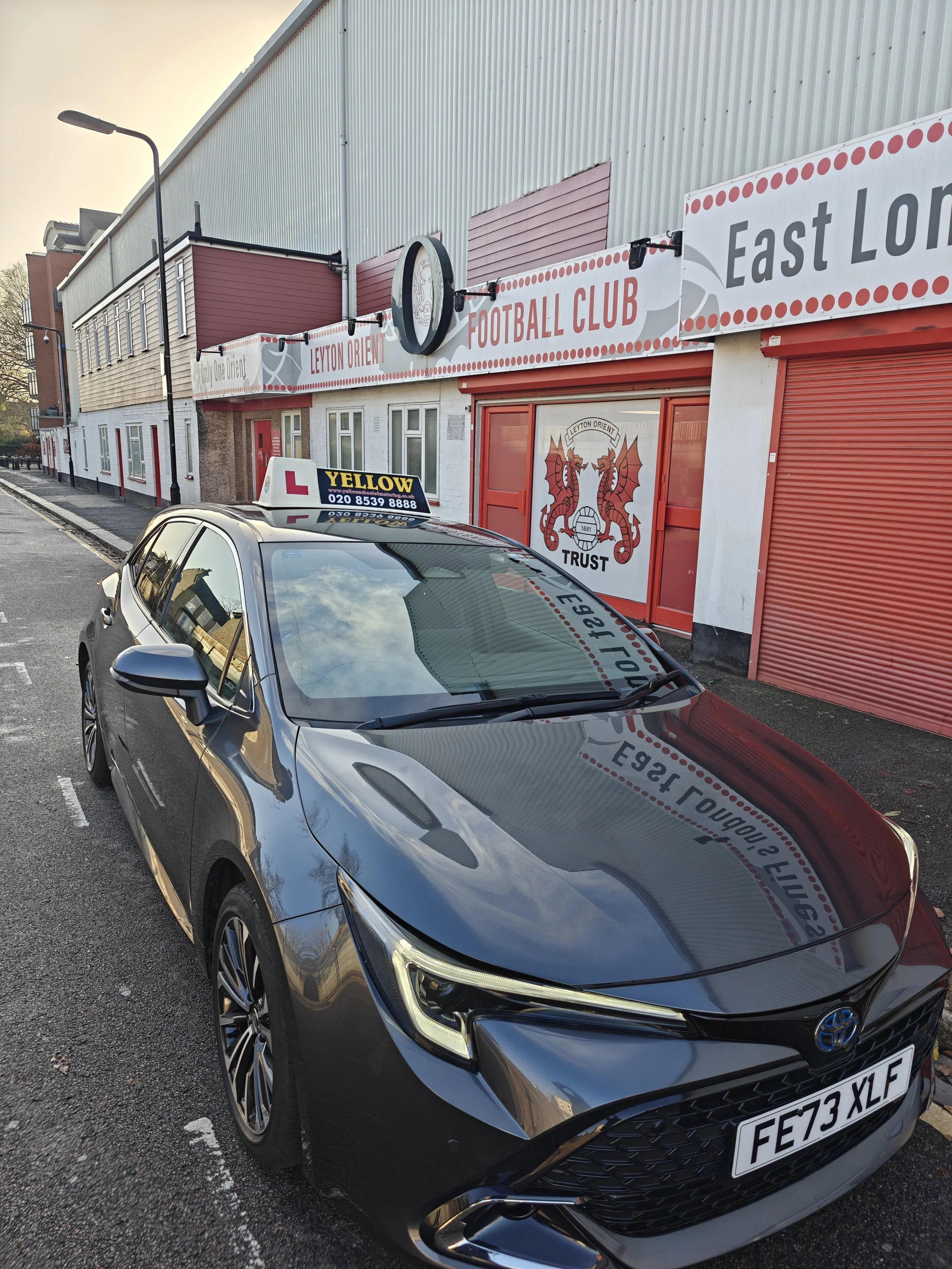 Image of a yellow school of motoring instructor car parked outside the iconic leyton orient stadium waiting to pick up a student for a automatic lesson in the dual controlled car
