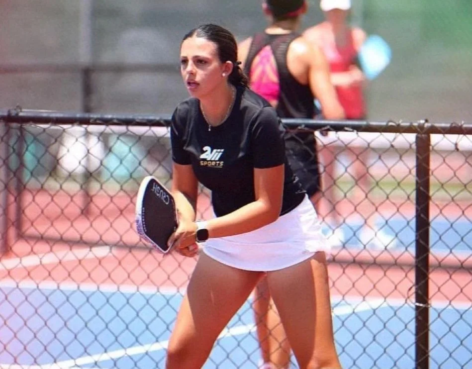 Juvenil mujer jugando al pickleball en la cancha exterior con una raqueta, usando uniforme deportivo, y una red de malla metálica.