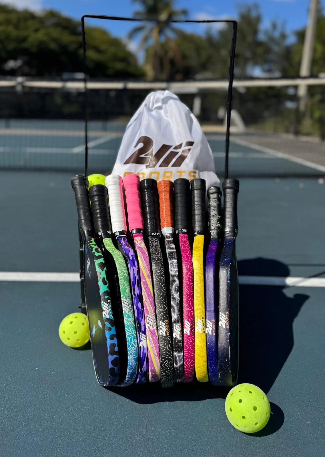 Raqueta de tennis con varias raquetas en diferentes colores y dos pelotas de tenis en una cancha de tenis al aire libre.