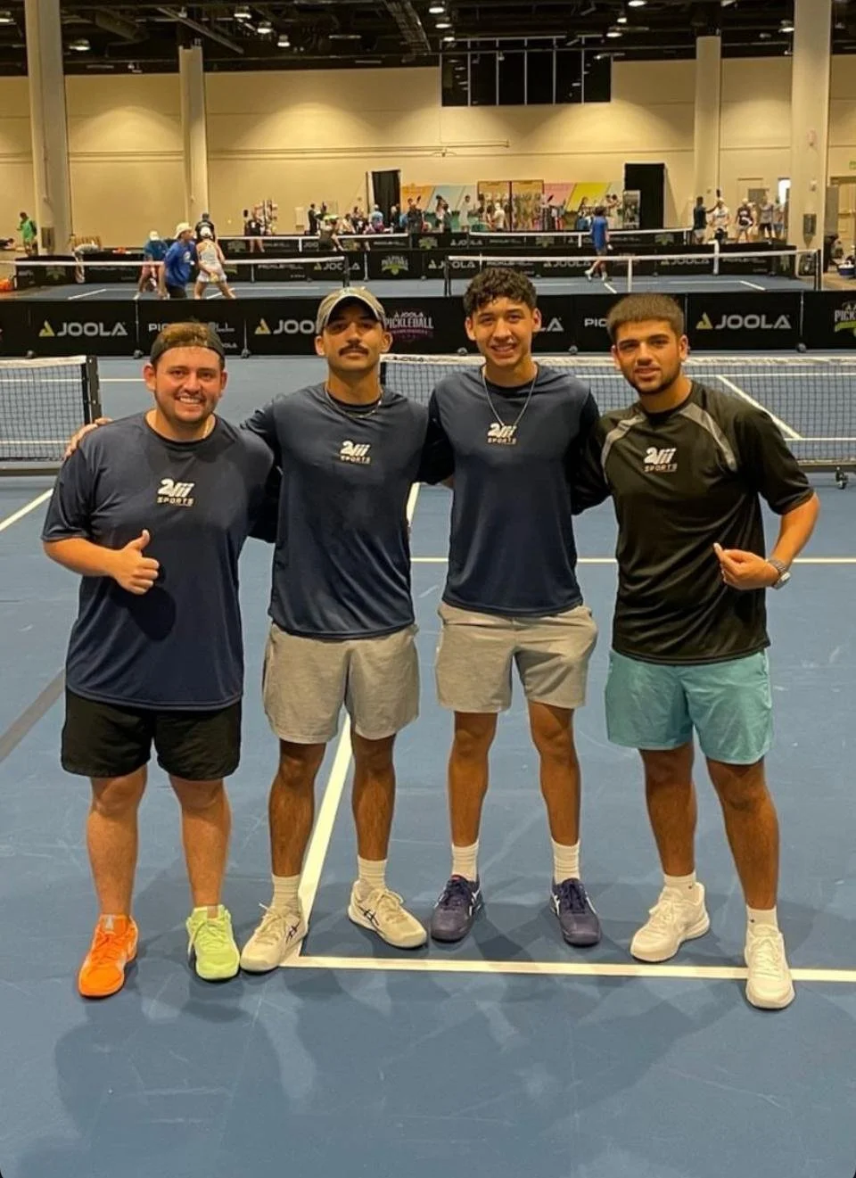 Cuatro hombres de pie en una cancha de pickleball en un evento o torneo, con varias mesas de pickleball y otras personas jugando en el fondo.