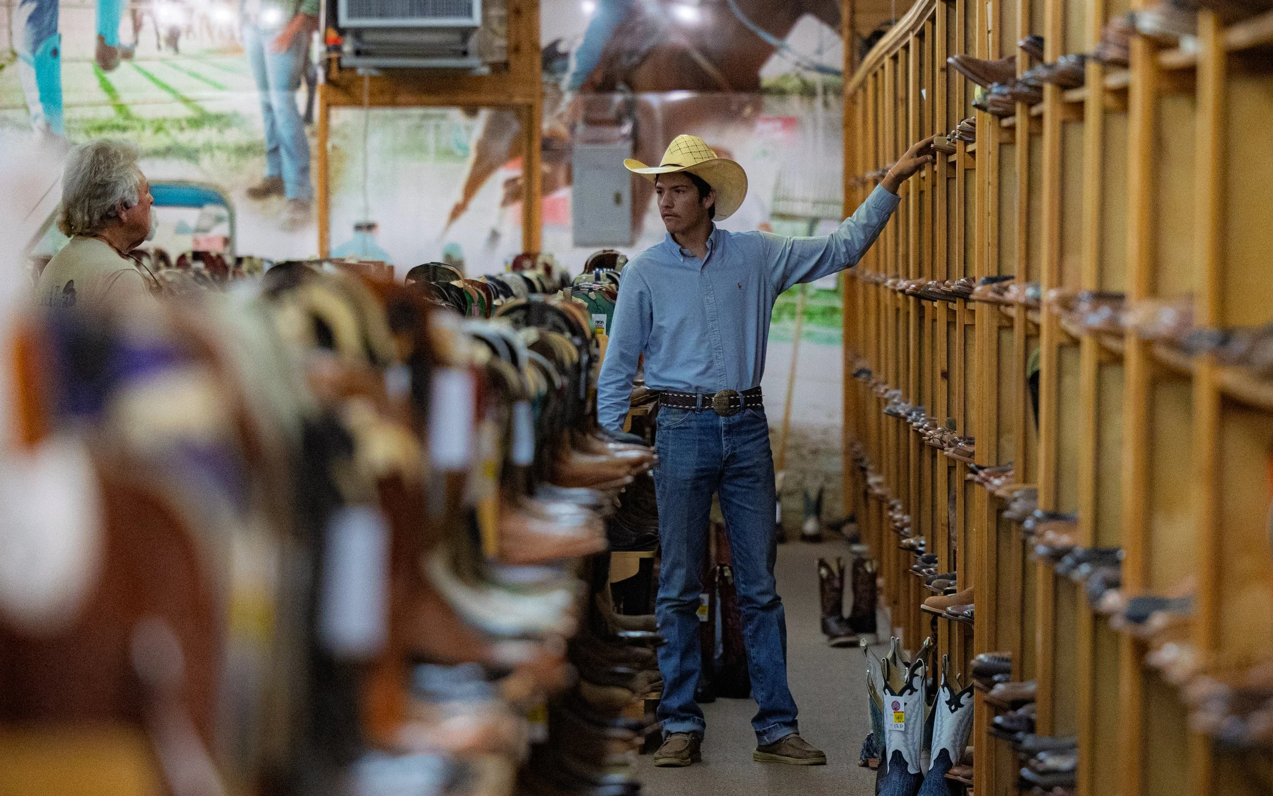 Young man wearing a cowboy hat and a light blue shirt shopping for cowboy boots at an outdoor or semi-outdoor venue, with shelves filled with various pairs of boots.