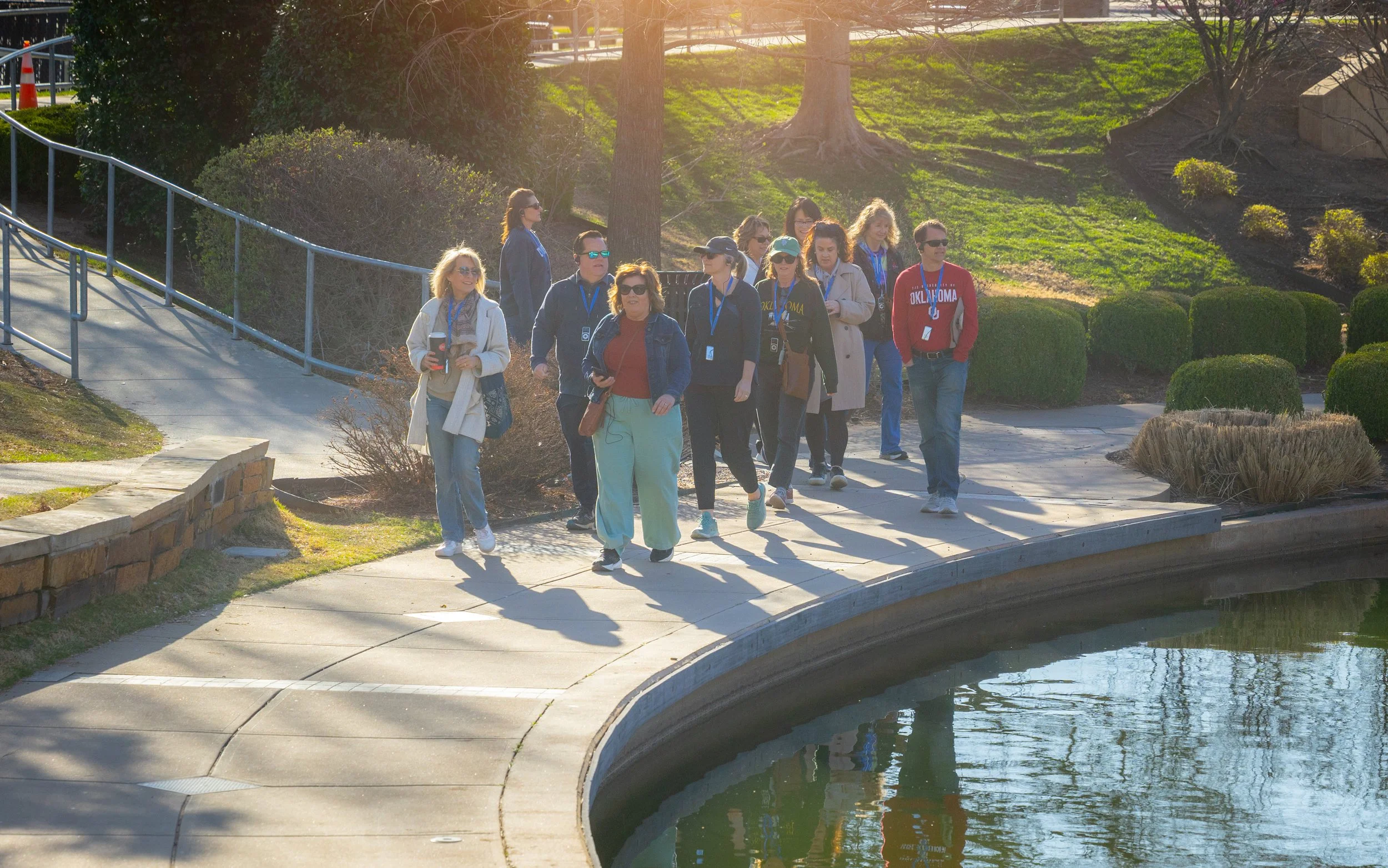 A group of people walking along a paved path next to a water feature in a park, with trees and bushes in the background, during daytime with sunlight casting shadows.