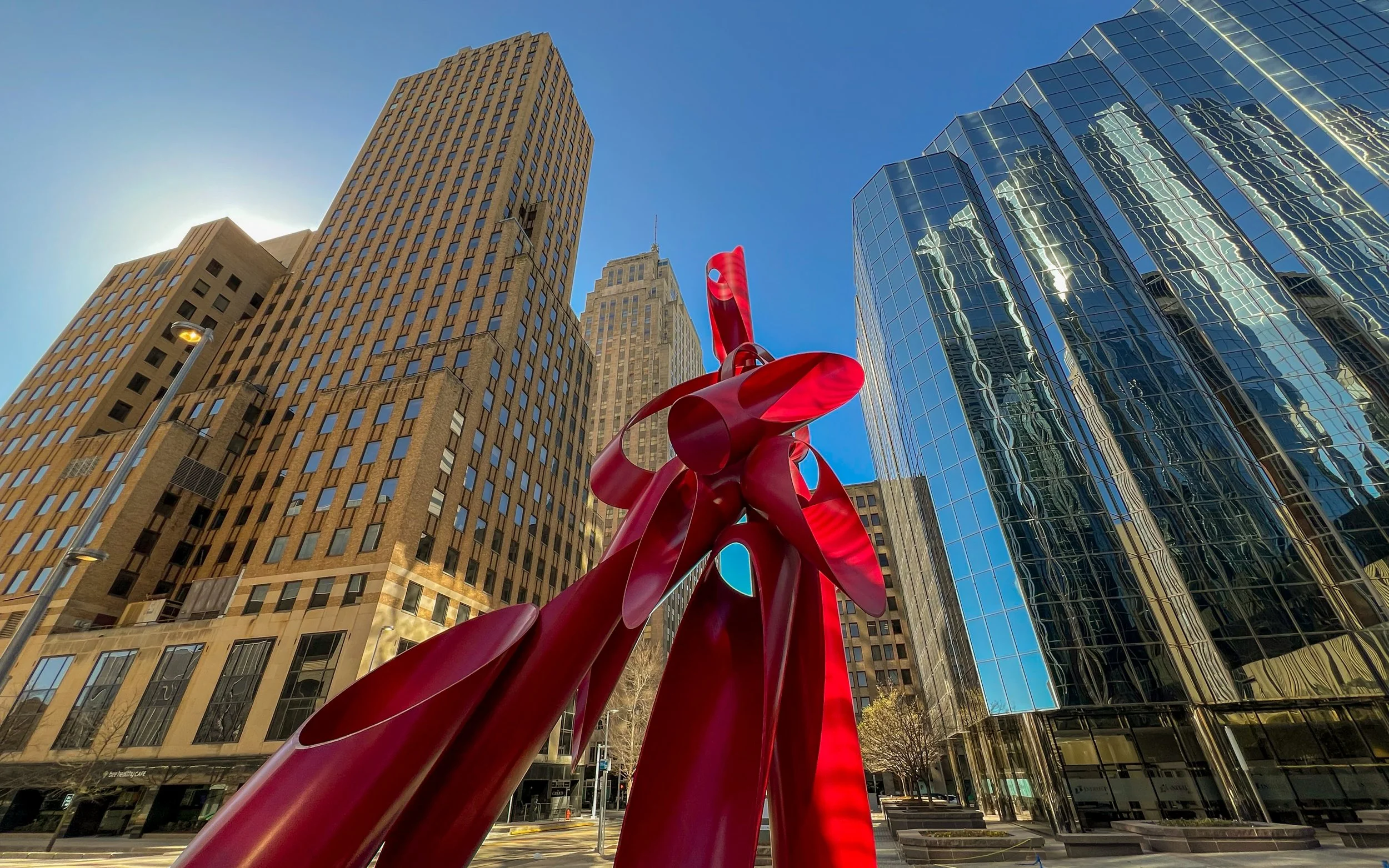 A tall, abstract red sculpture in front of modern glass and brick buildings in an urban downtown area.
