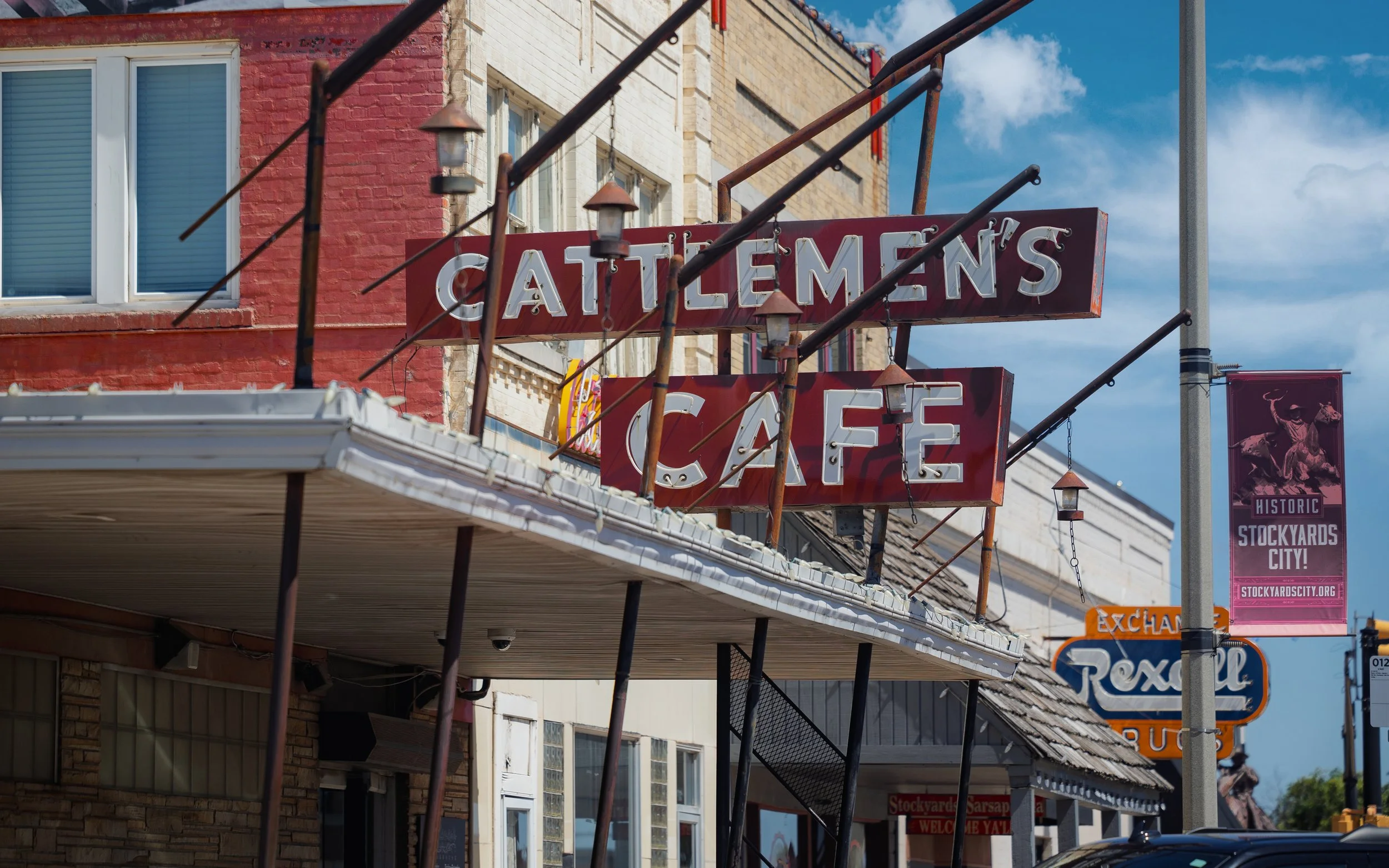 Vintage sign for Cattlemen's Cafe, with rusted metal and illuminated letters, on a building in a historic district, under a partly cloudy sky.