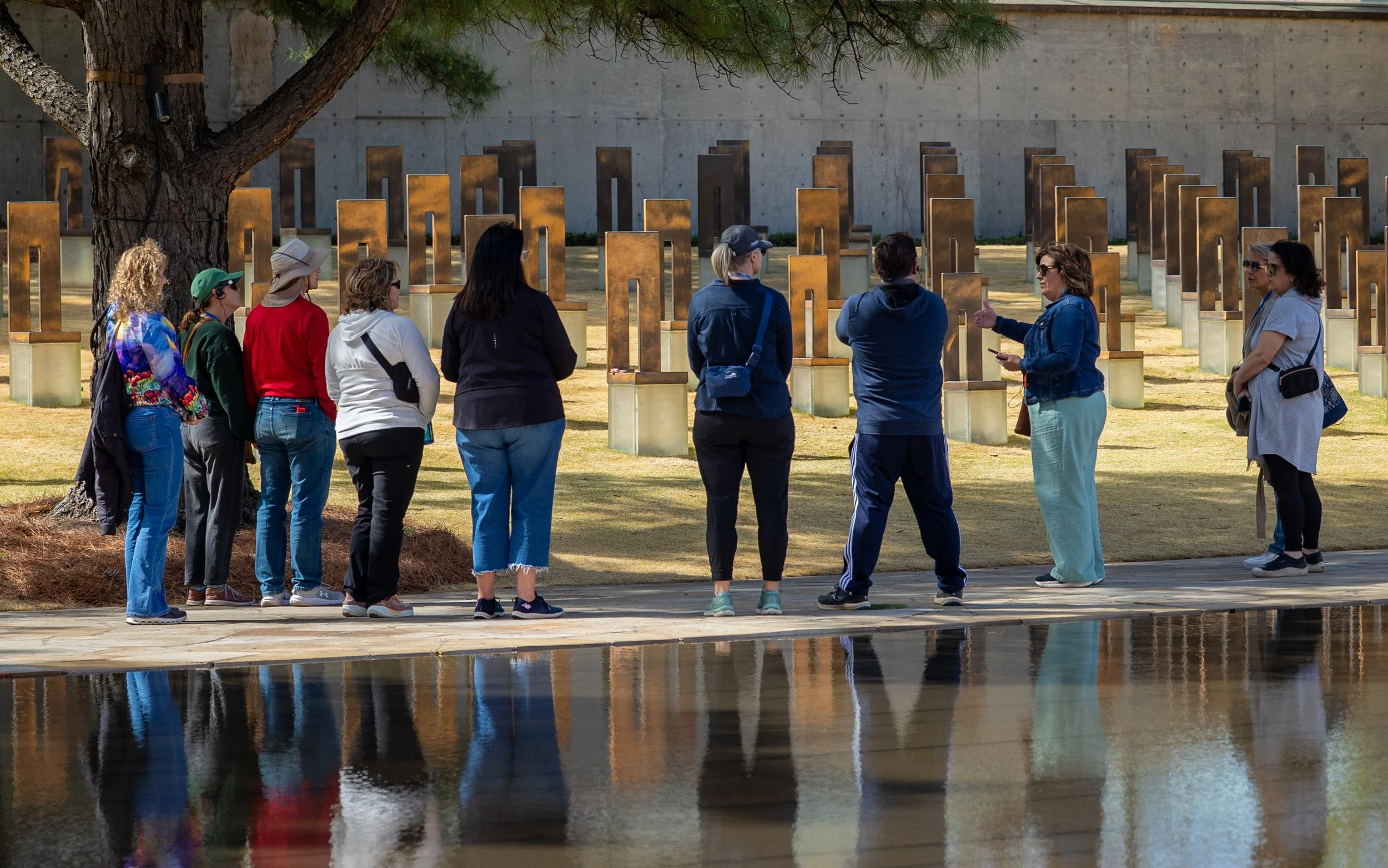 Group of people visiting a memorial with rectangular metal plaques, standing on a paved walkway near a water feature, with a large tree and concrete wall in the background.