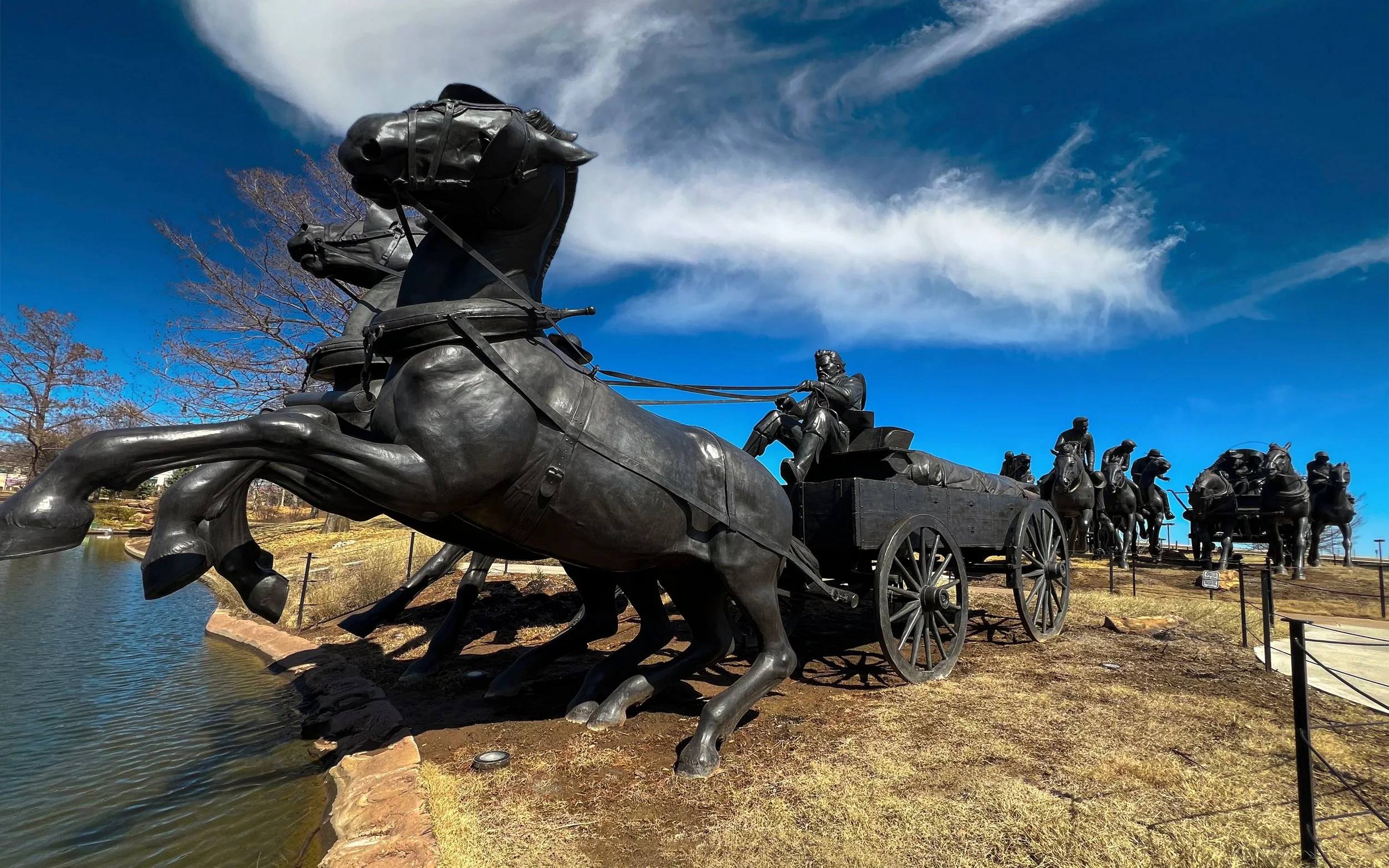 Statue of a horse-drawn carriage with multiple horses and drivers, located outdoors near a body of water, under a cloudy blue sky.