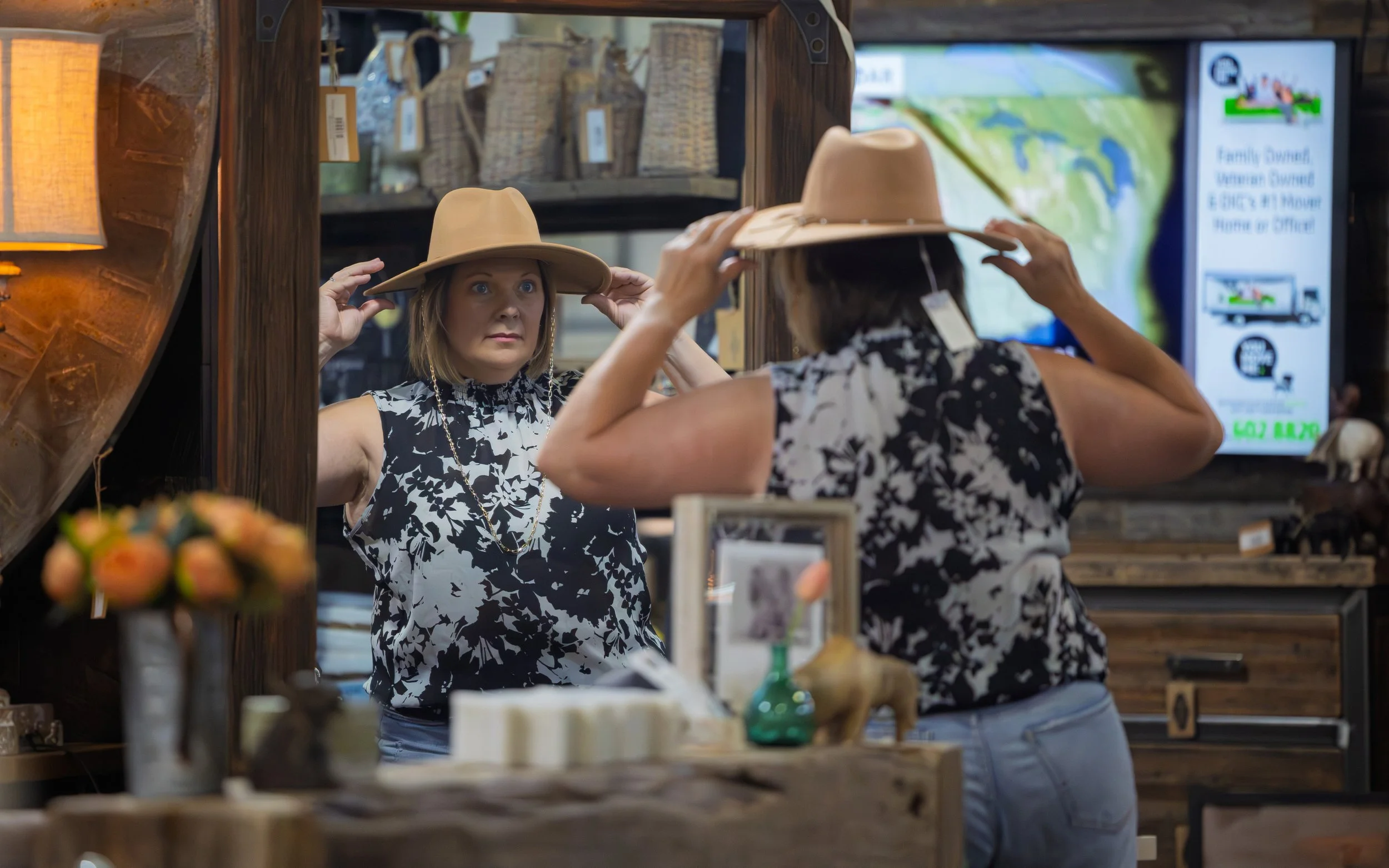 A woman trying on a tan hat in front of a mirror inside a store, with various decorative items and a digital display in the background.