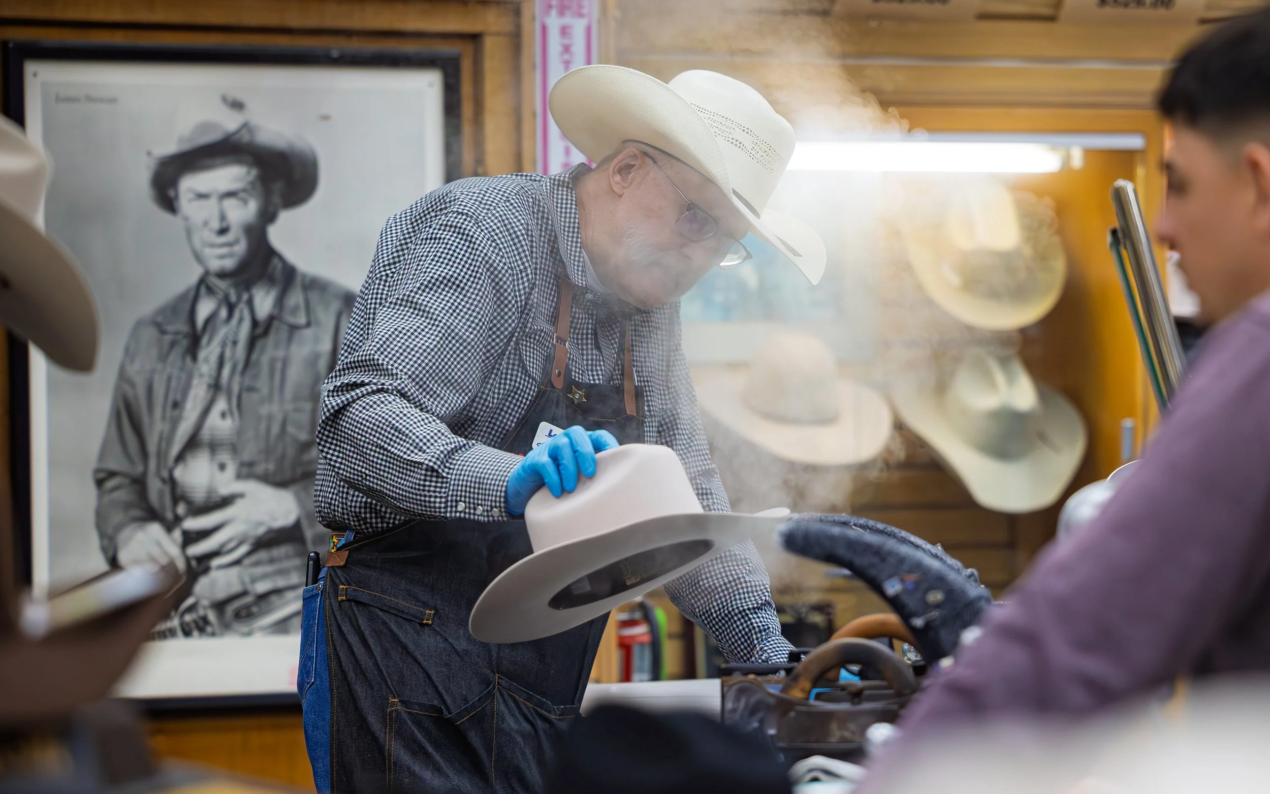 An older man wearing a cowboy hat, glasses, a checkered shirt, and blue gloves is working with a hat in a hat shop. There are other hats on display on the wooden wall and a black-and-white portrait of a man in a cowboy hat in the background.
