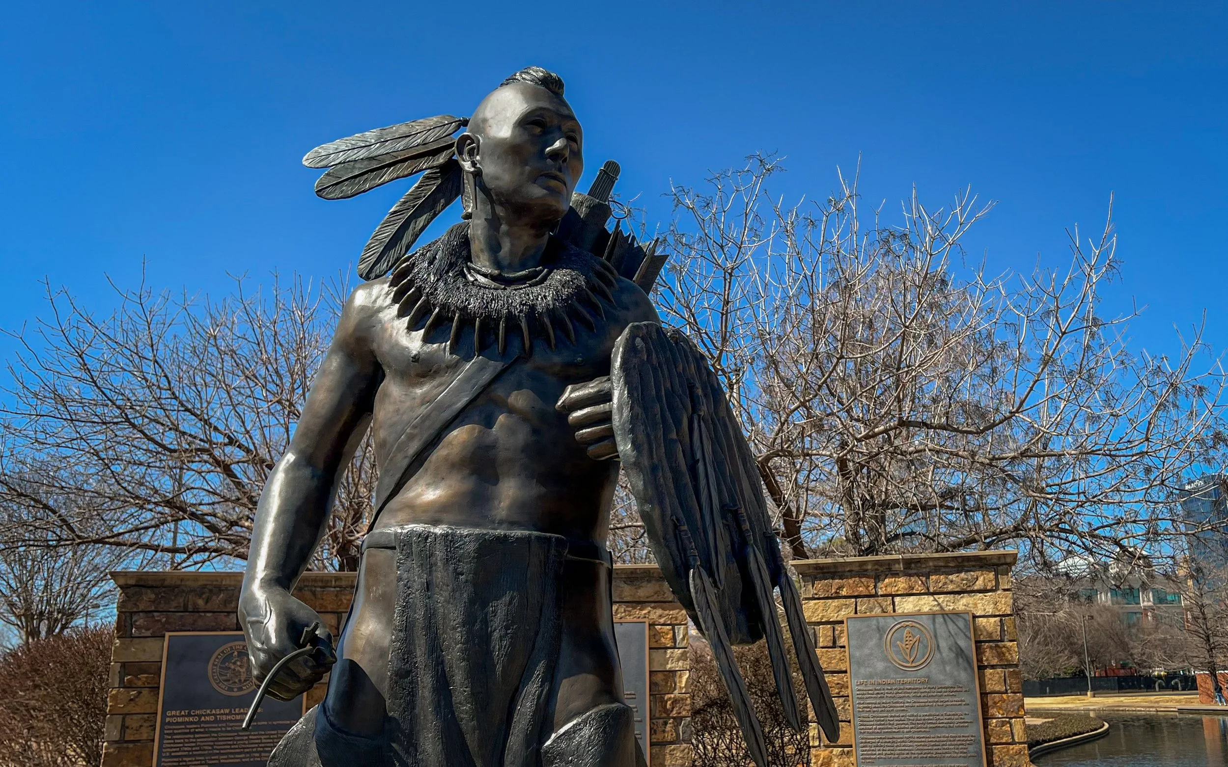 Bronze statue of a Native American man with large feathers in his hair, holding a shield in his left hand, set outdoors with leafless trees and stone plaques in the background.