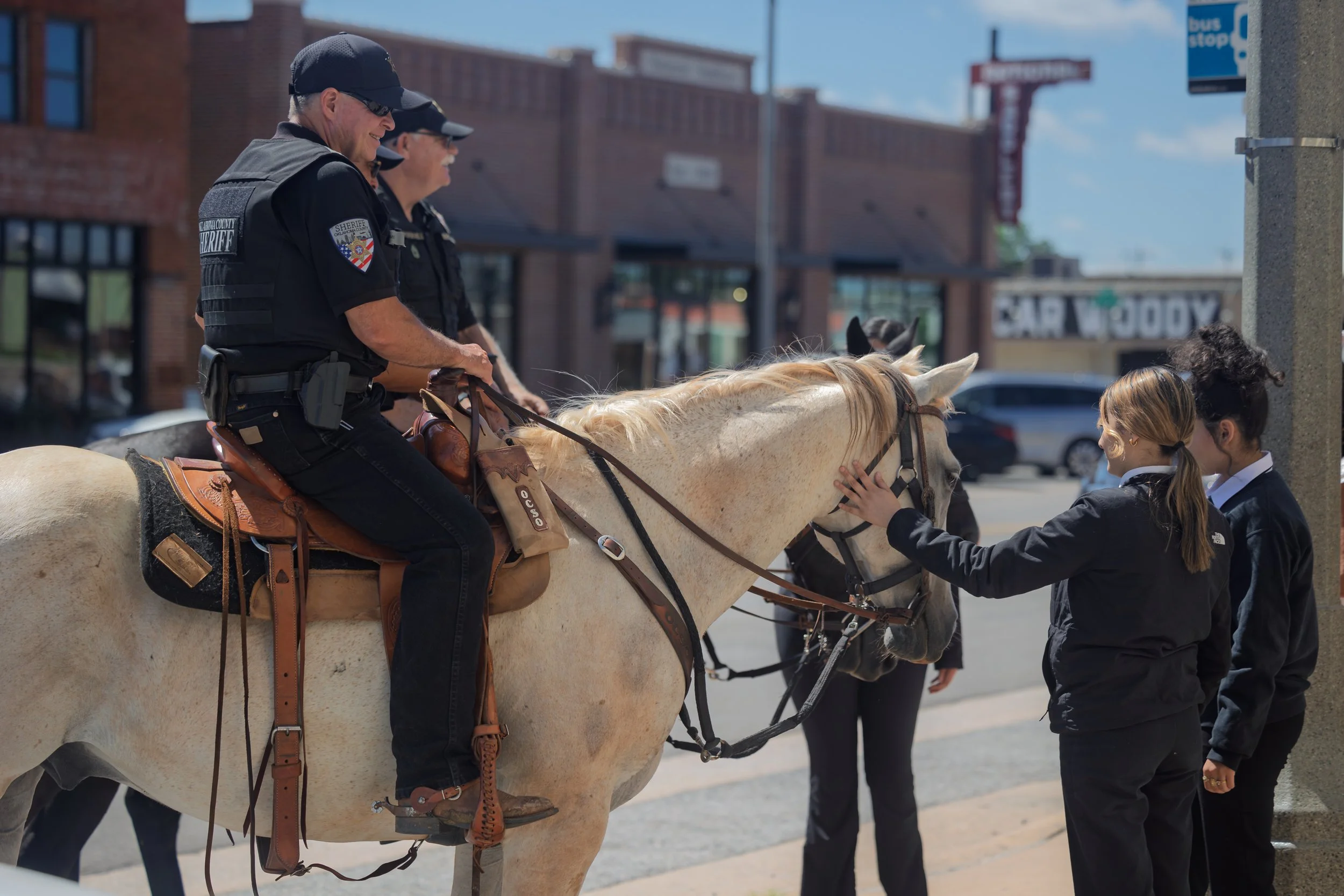 Two police officers on horseback interacting with two young women on a city sidewalk. The women are petting the horse.