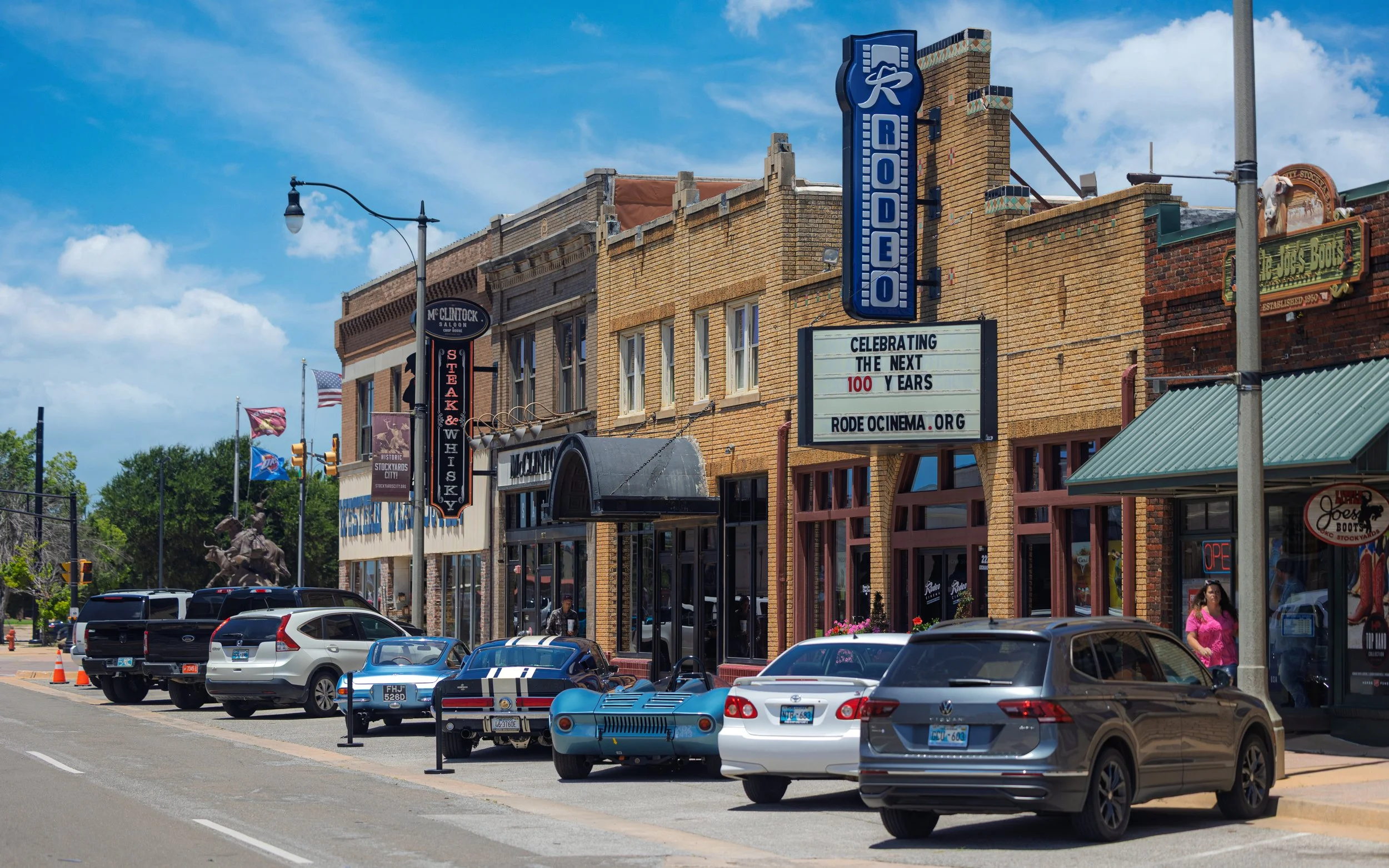 Street view of downtown with parked cars, storefronts, and signage on a sunny day with blue sky and scattered clouds.