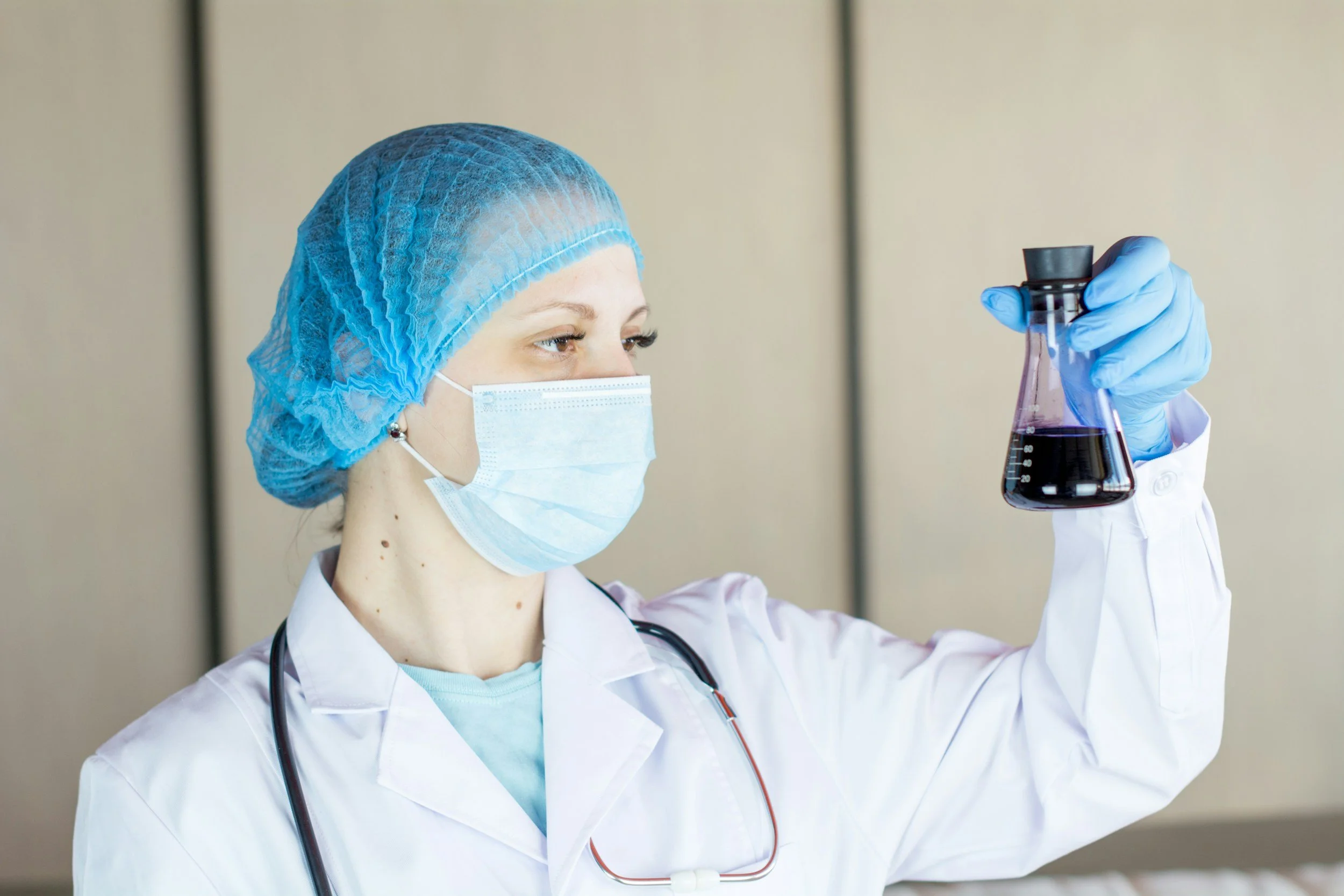 A female medical professional wearing a blue hairnet, face mask, and white lab coat, examining a test tube containing a dark liquid.