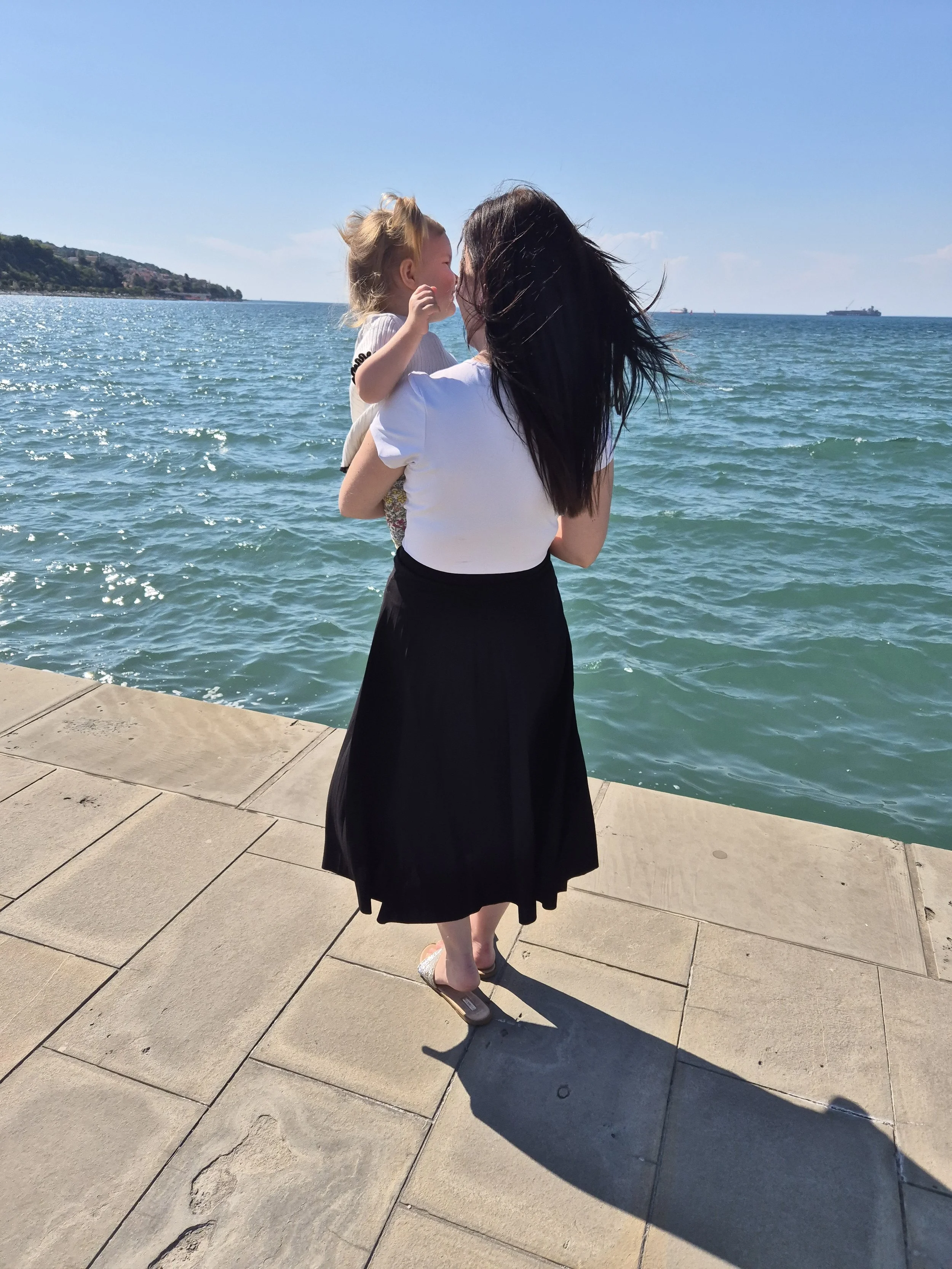 A woman with long dark hair in a white top and black skirt holding a young girl with blonde hair near a body of water on a sunny day.