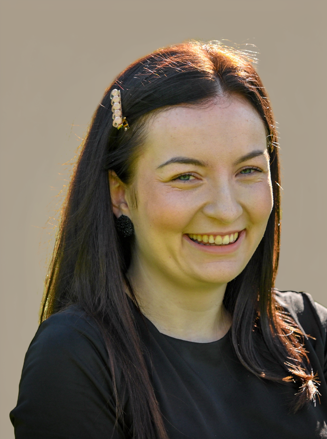 A smiling young woman with dark straight hair, a hair clip, black earrings, and a black top, outdoors in natural sunlight.