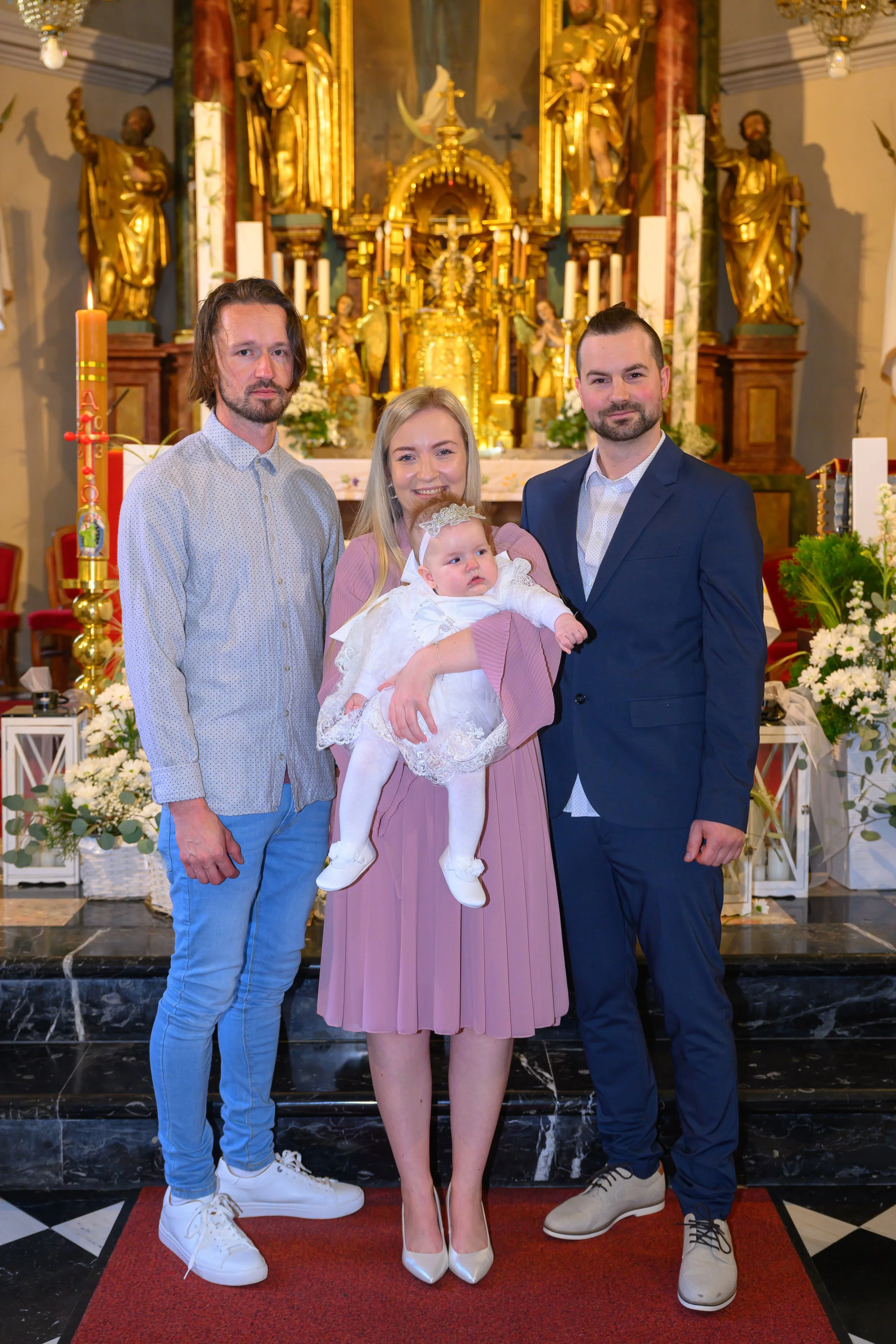 A family of four standing inside a church near an altar decorated with gold statues and religious icons. The woman in the middle is holding a baby girl dressed in white. The two men on either side are dressed in formal attire. All family members are posing for the photo.