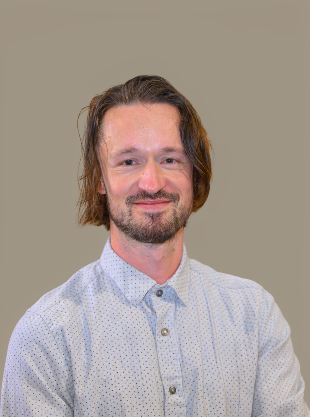 A man with shoulder-length brown hair and a beard, wearing a white button-up shirt with small polka dots, smiling against a plain beige background.