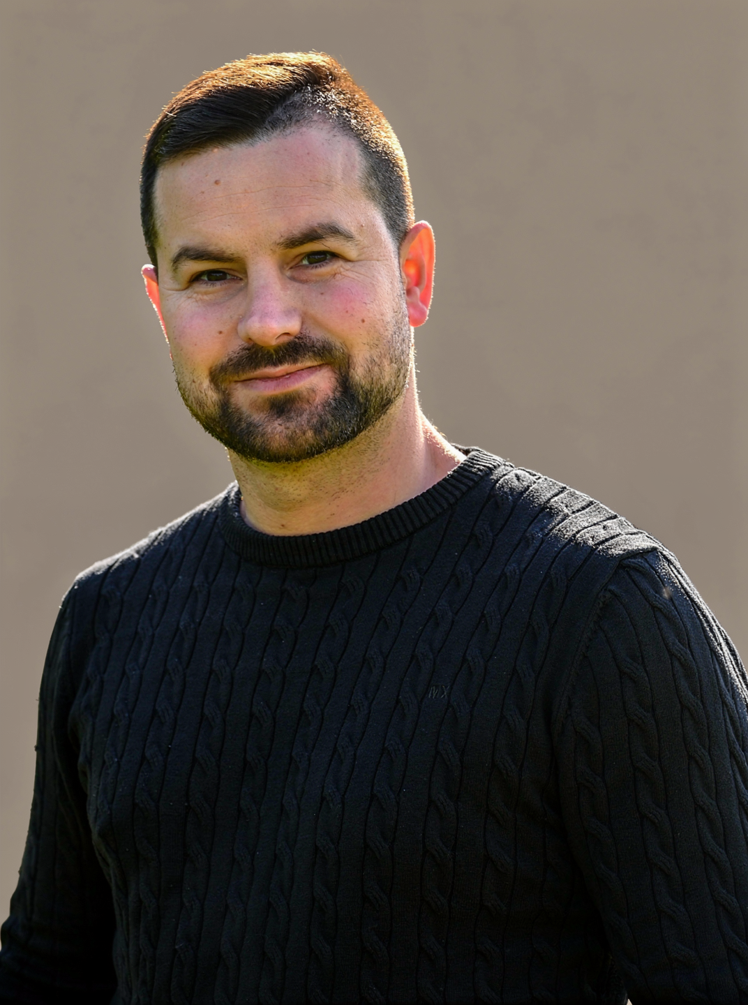 A man with short dark hair, a beard, and a slight smile wearing a black cable-knit sweater outdoors.