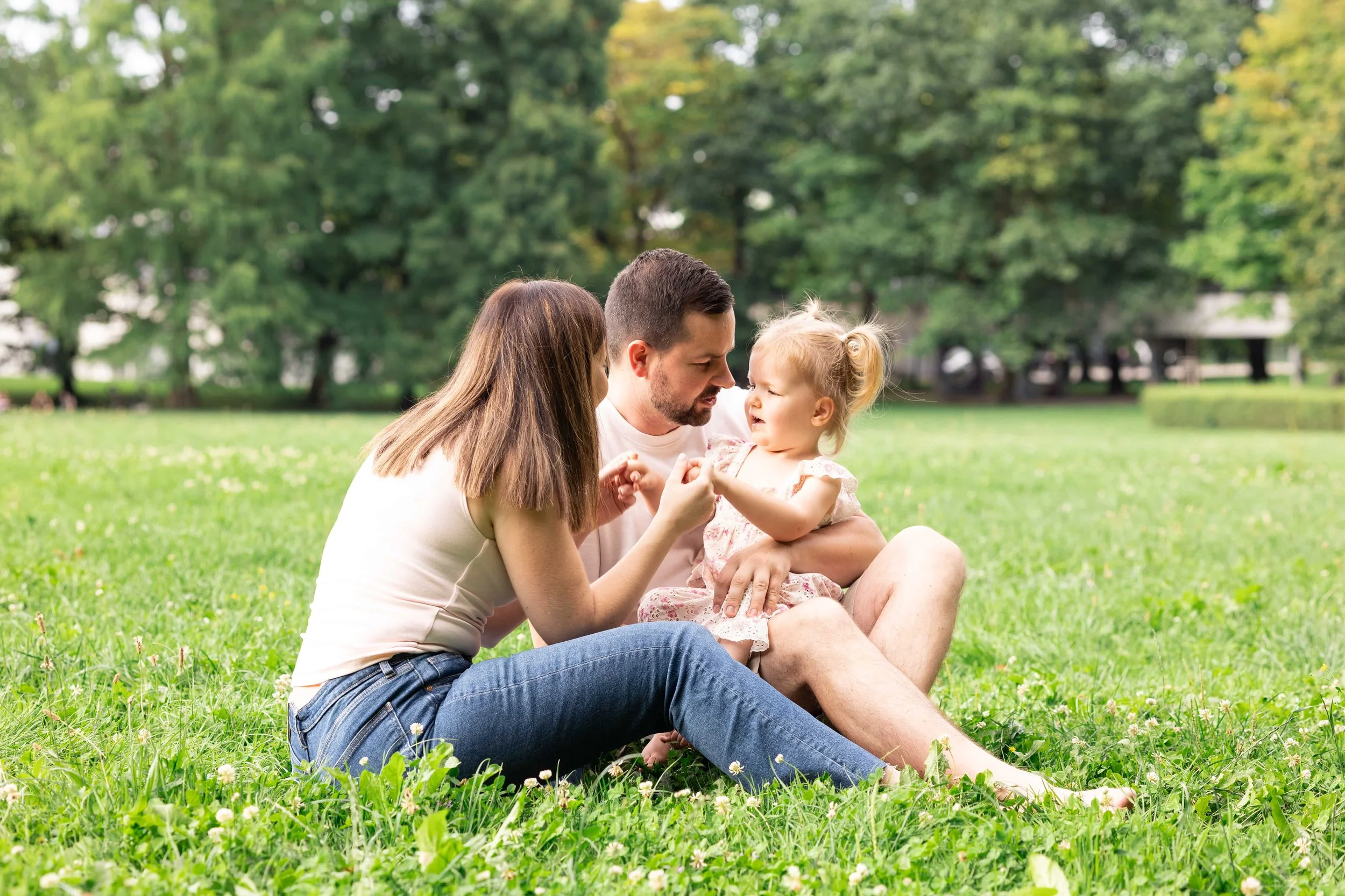 Family sitting on grass in a park, with trees in the background. The father, mother, and young girl are engaged in a close moment, possibly playing or talking.