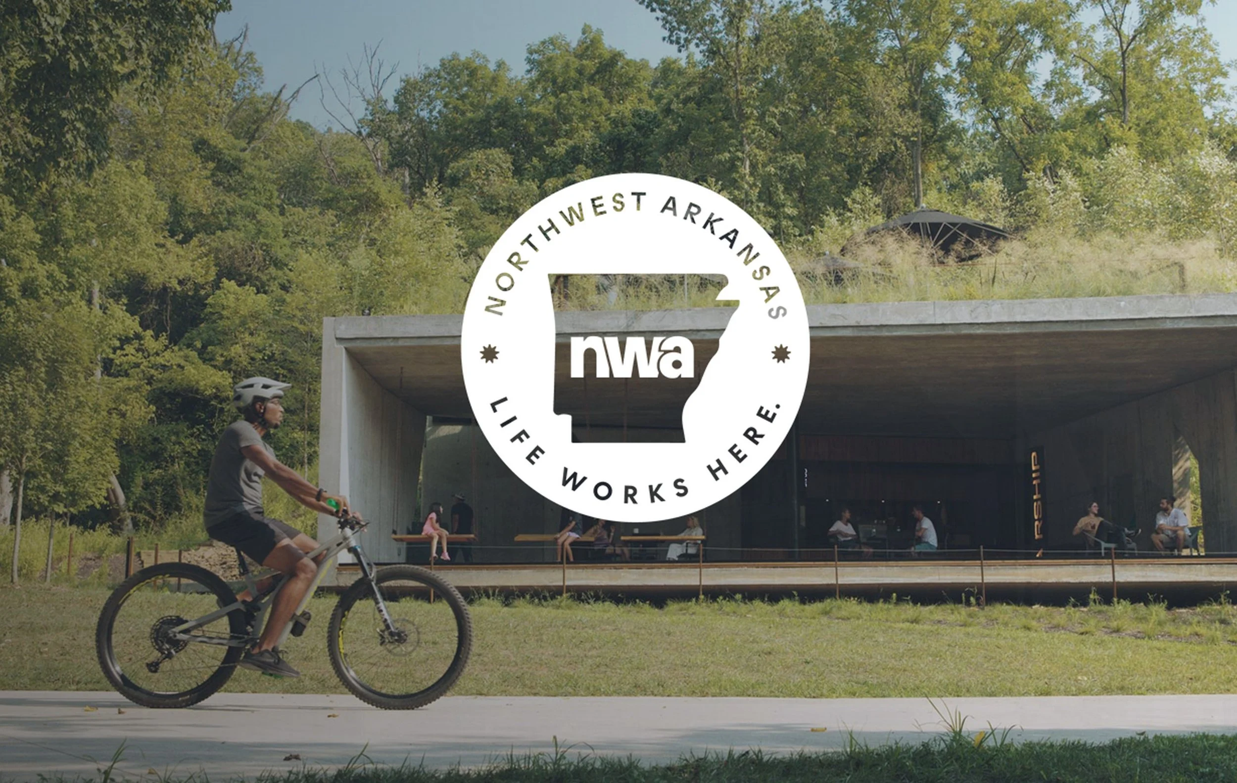 A person riding a bicycle past a modern building with a large open porch, surrounded by trees, with a logo overlay saying "Northwest Arkansas Life Works Here. NWA".