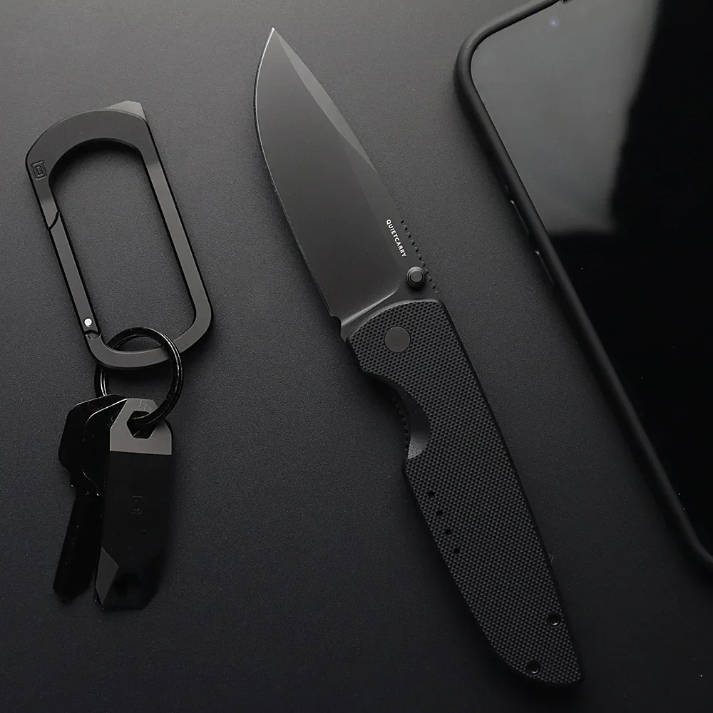 A black Out of Duty folding knife with textured handle, a black carabiner, and a set of keys on a black surface, with part of a smartphone visible.