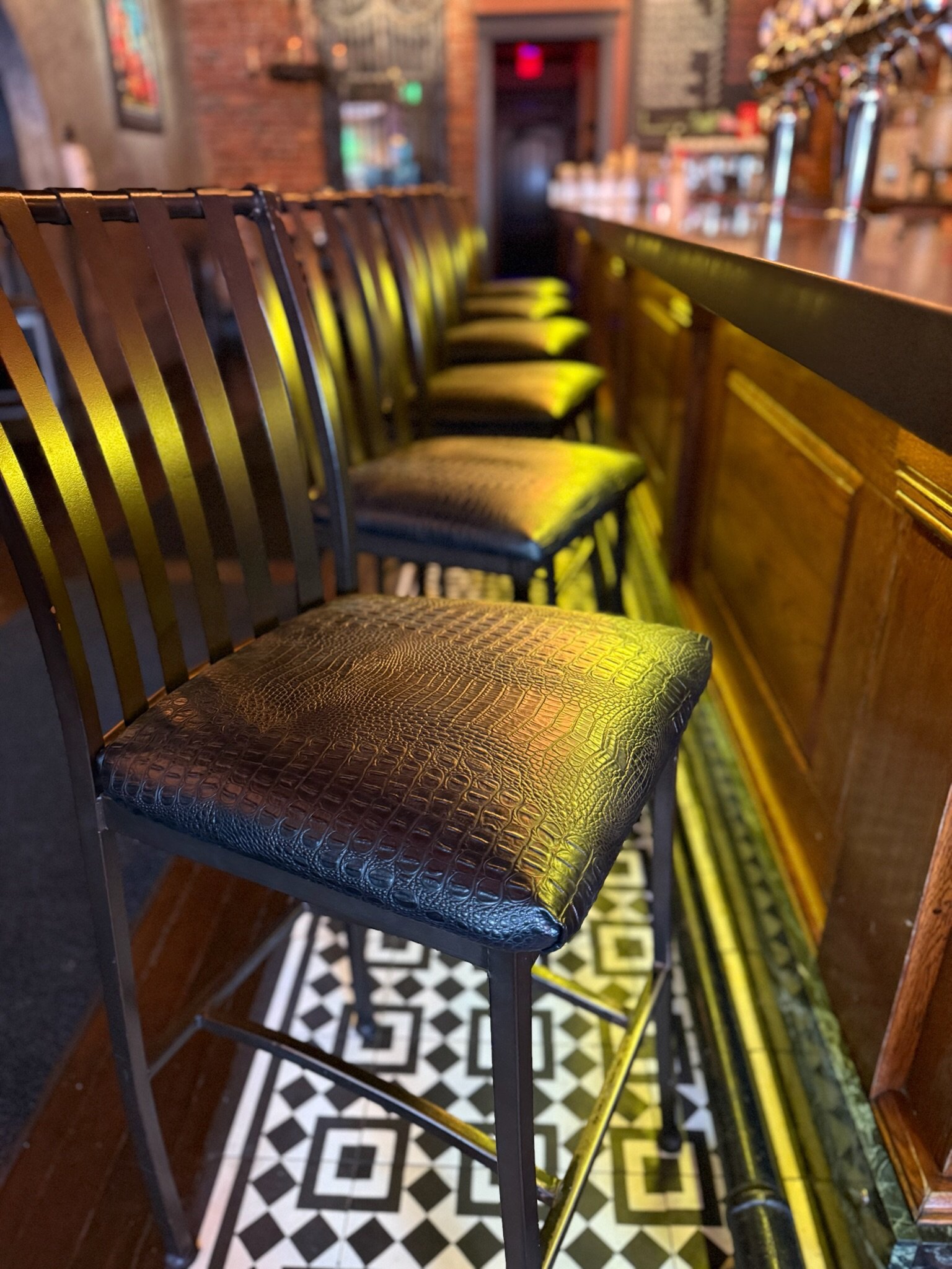 A row of black bar stools with leather seats at a bar in a restaurant with patterned black and white tile floor.