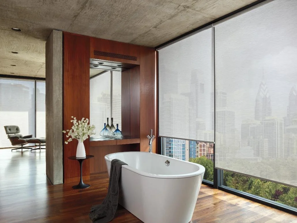Modern bathroom with a white bathtub, large windows with city view and blinds, wooden and concrete walls, a black side table with a flower vase, and decorative bottles, in an urban apartment.