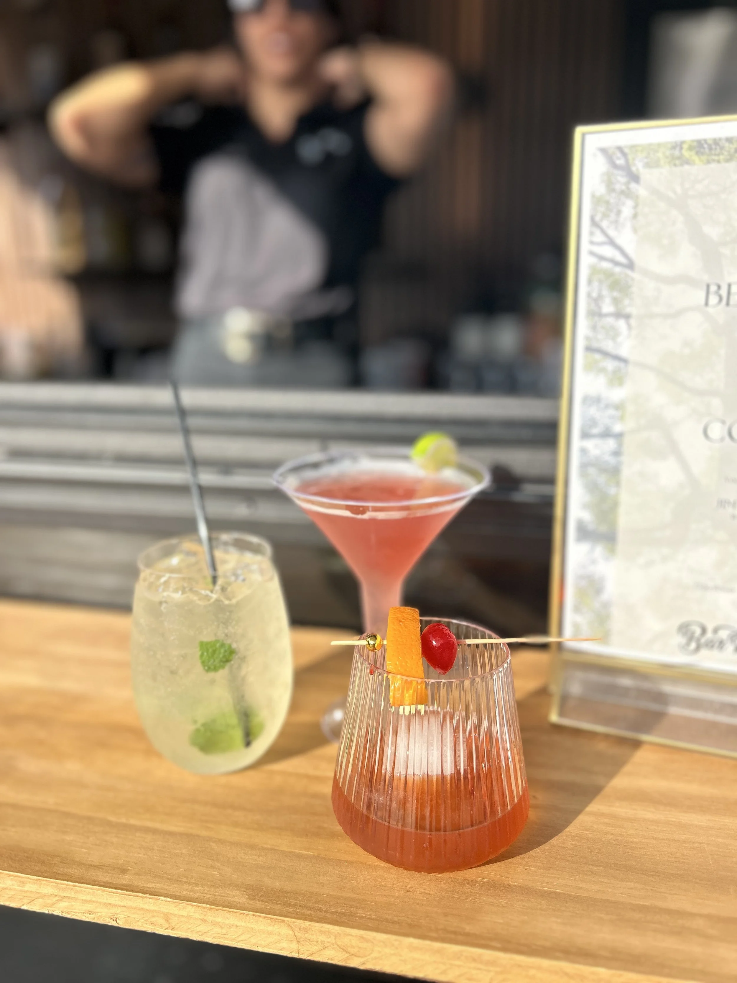 Bar Rouler mobile bar serving three colorful cocktails on a wooden table with a blurred bartender and menu in the background.