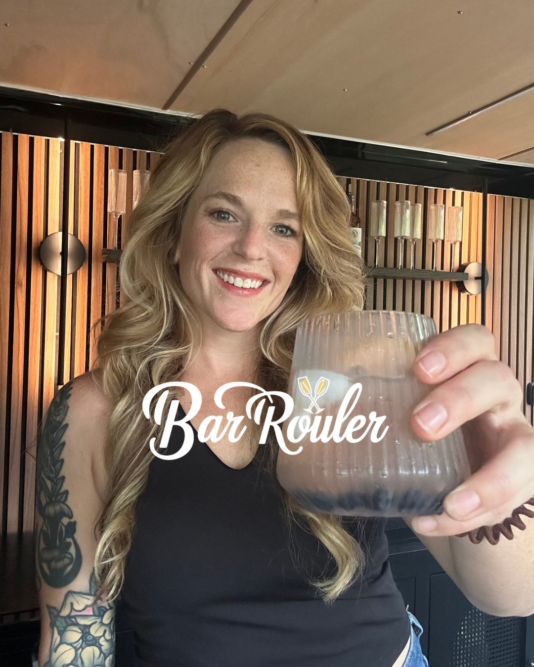 Bar Rouler serving cocktailsl. A smiling woman with wavy blonde hair holding a drink in a textured glass. She has tattoos on her arms and is wearing a black sleeveless top, standing in front of a polished mobile bar with slatted walls, light fixtures
