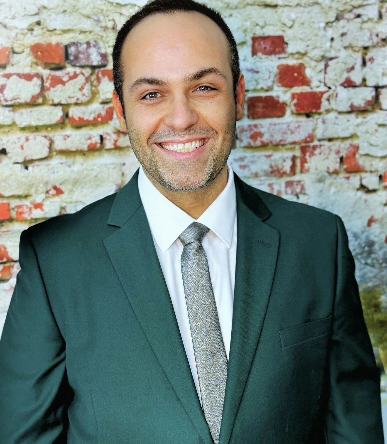 A smiling man in a dark suit and grey tie standing in front of a weathered brick wall.