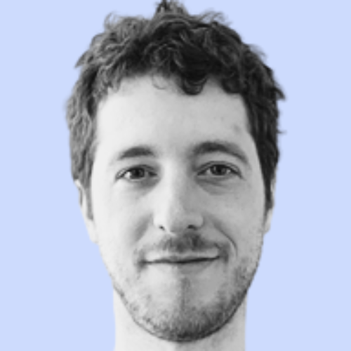 A close-up black and white portrait of a young man with curly hair, slight beard, and a subtle smile, set against a light blue background.