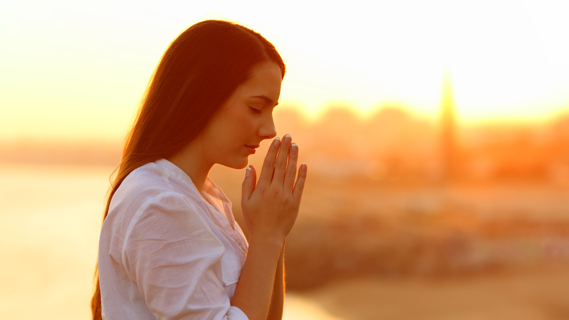 A woman with long brown hair in a white shirt is praying outdoors at sunset with her eyes closed and hands pressed together.