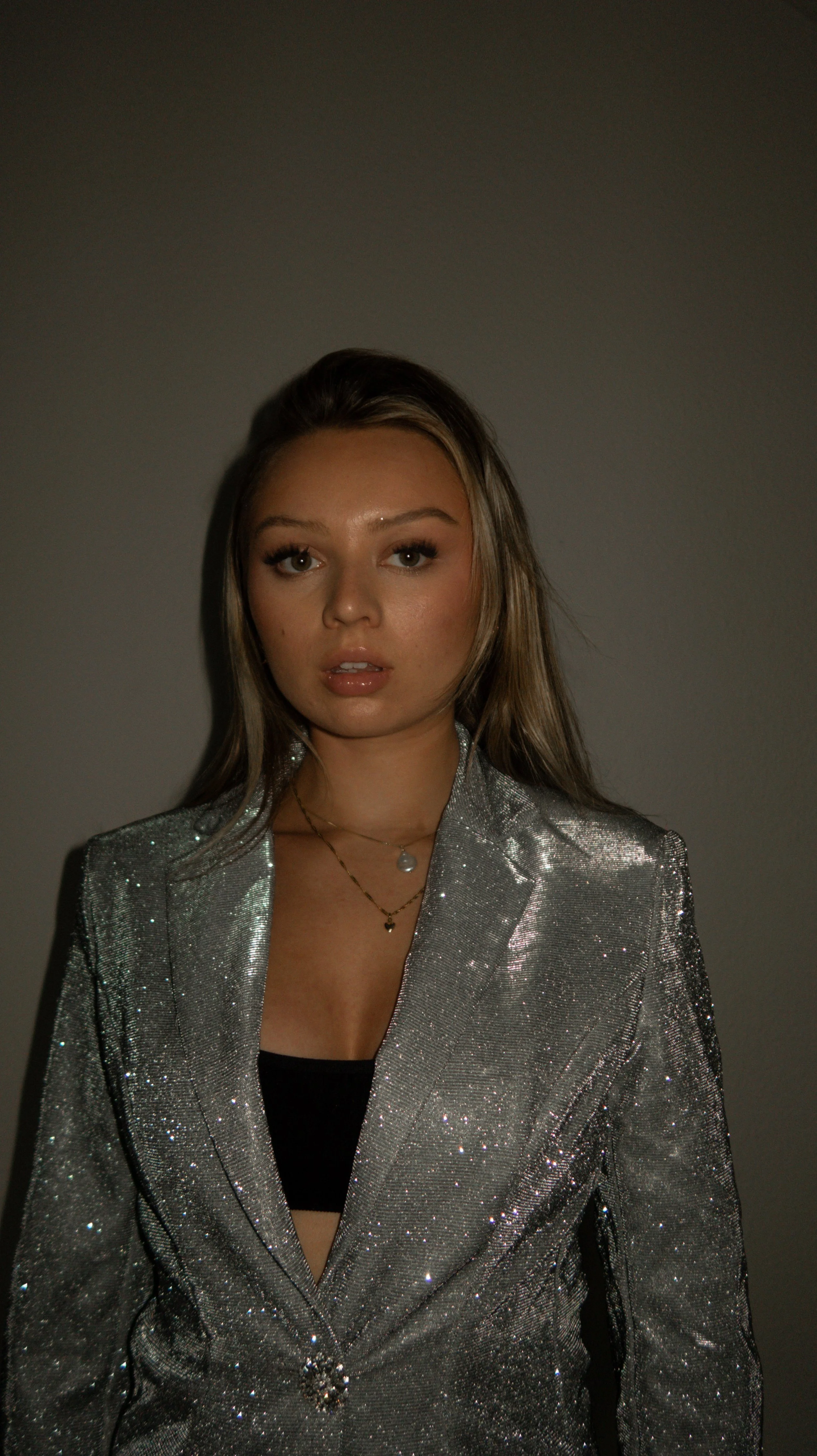 Young woman with long brown hair wearing a shiny silver blazer and layered necklaces standing against a gray background.