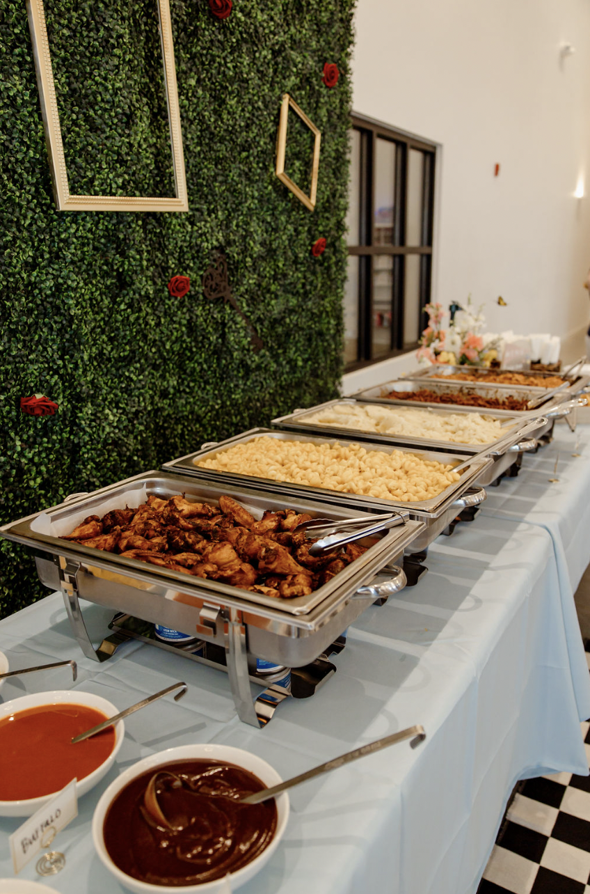 Buffet table with trays of chicken, mashed potatoes, and other dishes against a green wall with decorative empty frames and red roses.