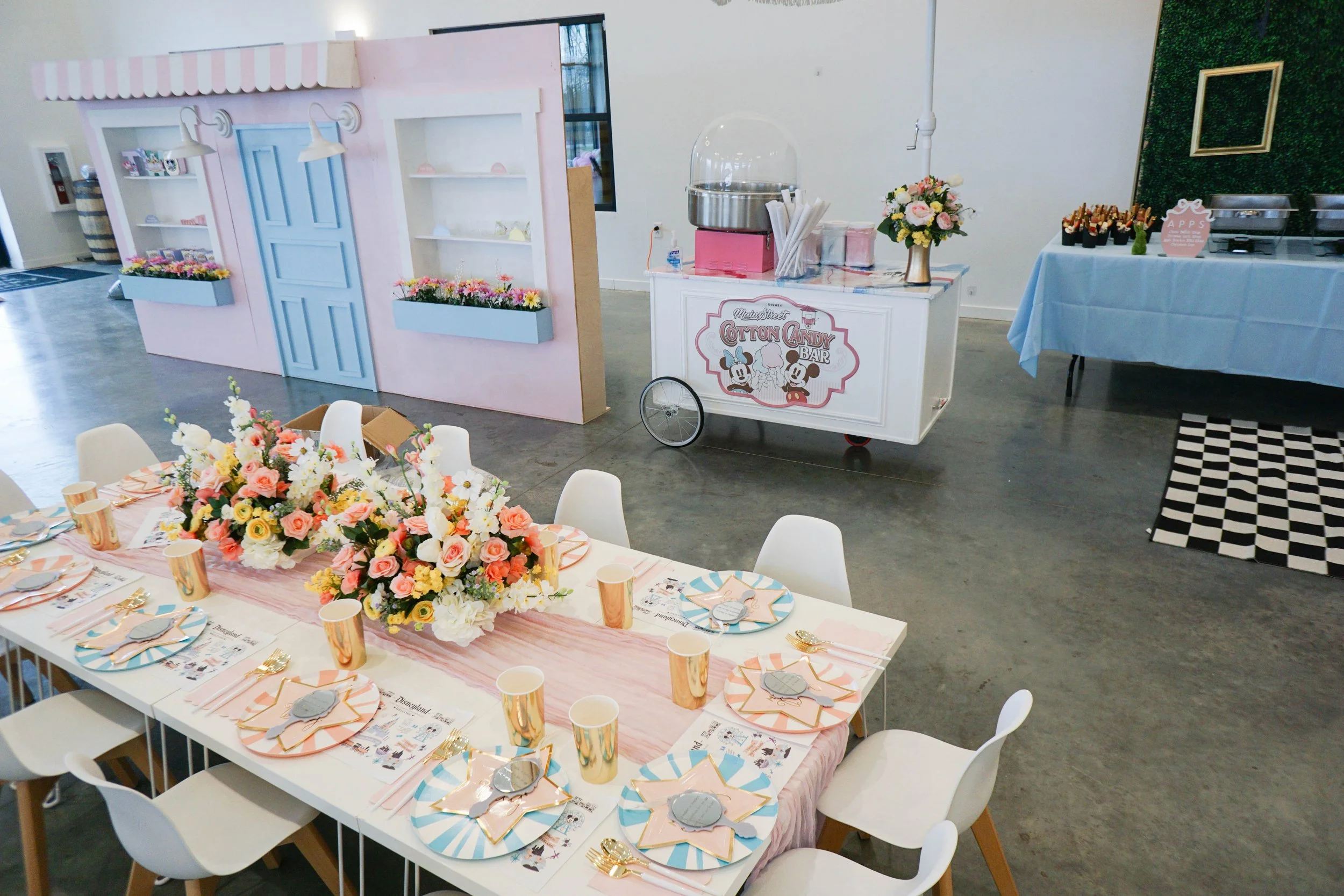 Decorated party space with a long table set with pink and blue striped plates, gold cups, and floral arrangements. In the background, a cotton candy stand with a Mickey Mouse logo, cupcakes, and a candy table.