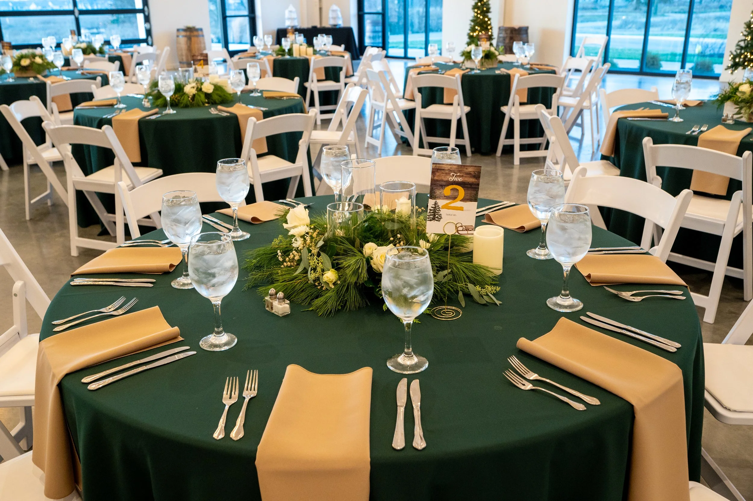 Round banquet table decorated with a green tablecloth, tan napkins, and glassware, with a floral centerpiece of white flowers and greenery, set up for a formal event in a well-lit room with large windows and Christmas trees in the background.