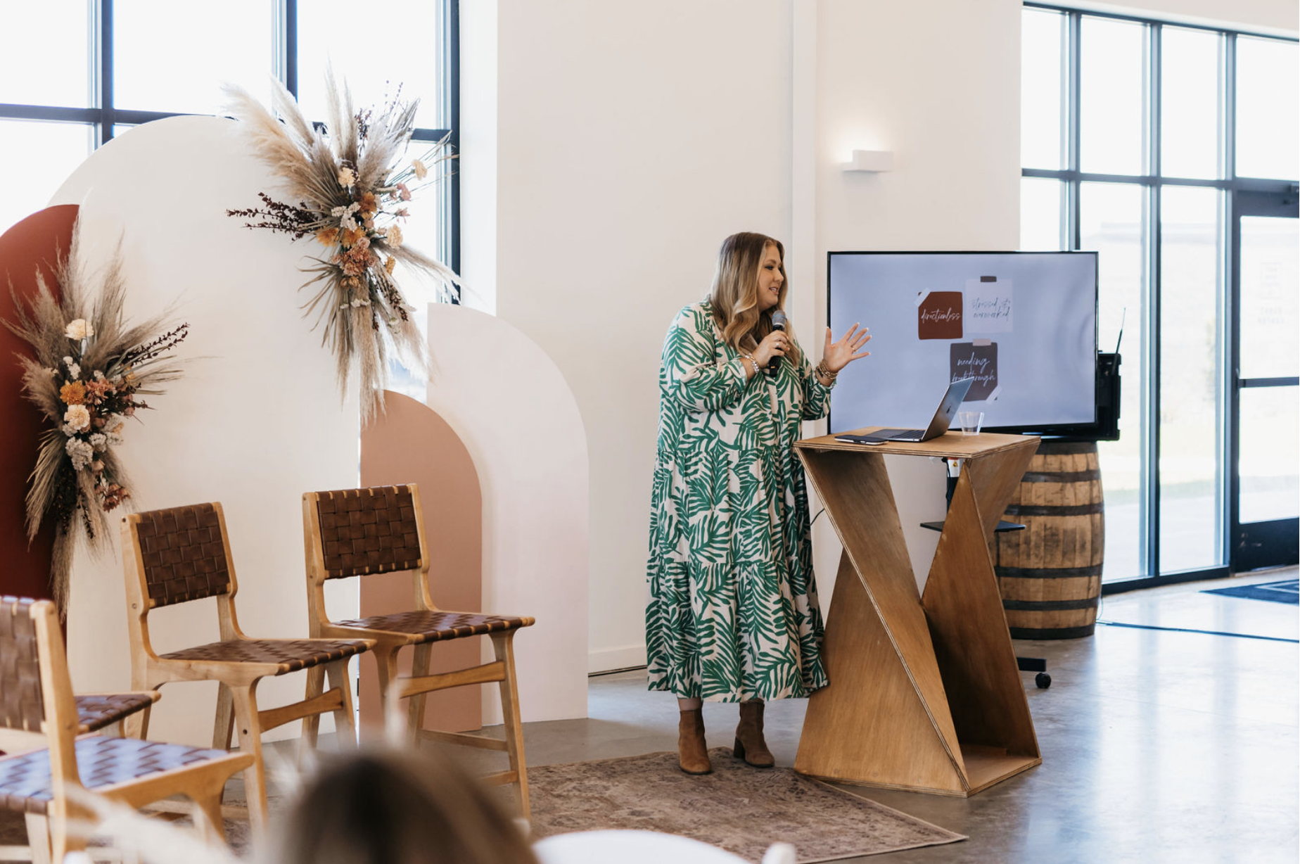 A woman in a green leafy patterned dress speaking into a microphone in a modern indoor space with large windows, a digital screen, wooden furniture, and decorative dried flowers on the wall.