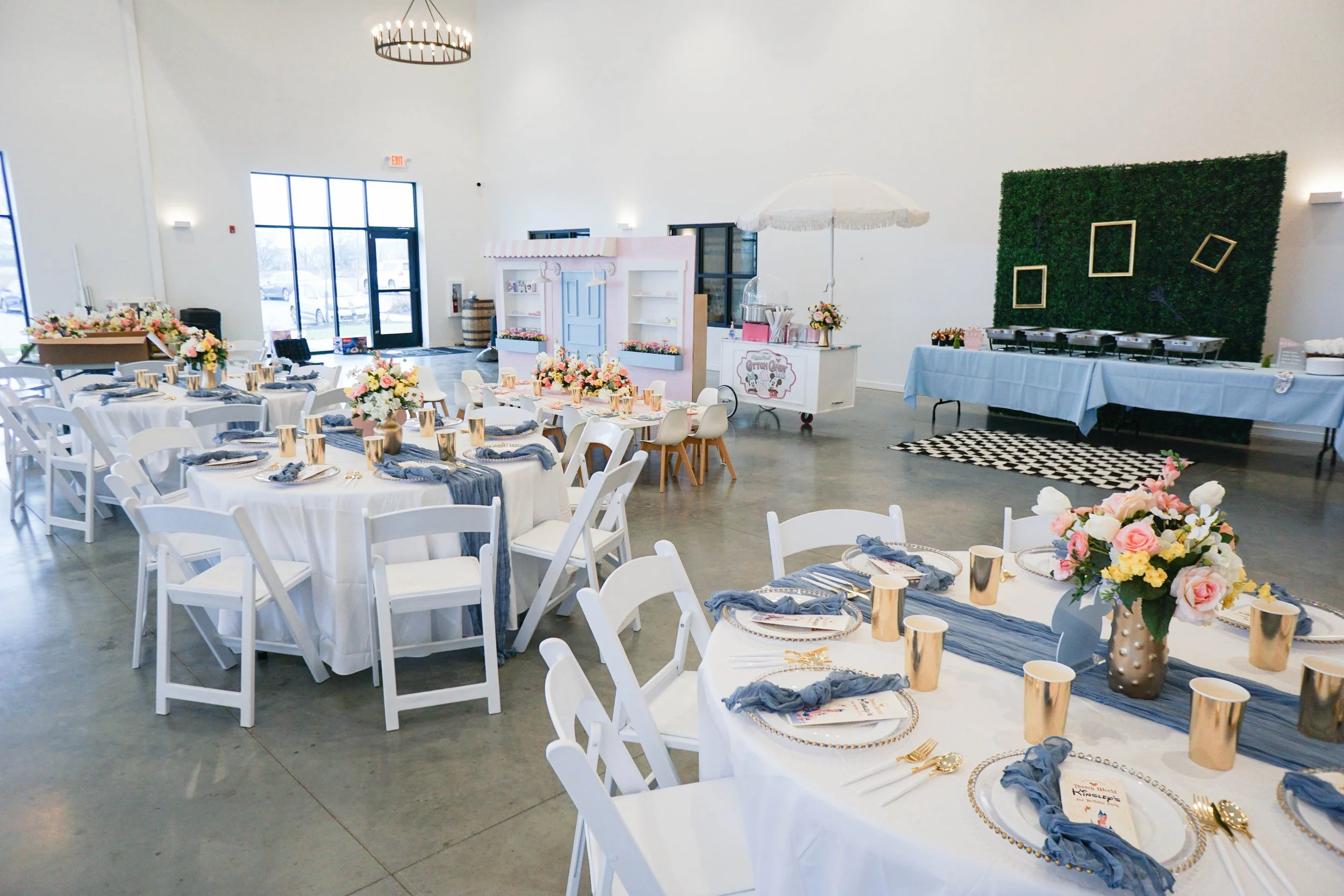 Elegant indoor event space decorated with round tables covered in white tablecloths, each with floral centerpieces, gold cowbell cups, and blue napkins. There are chait with white frames, a pink and white photo booth, a cotton candy station with an u