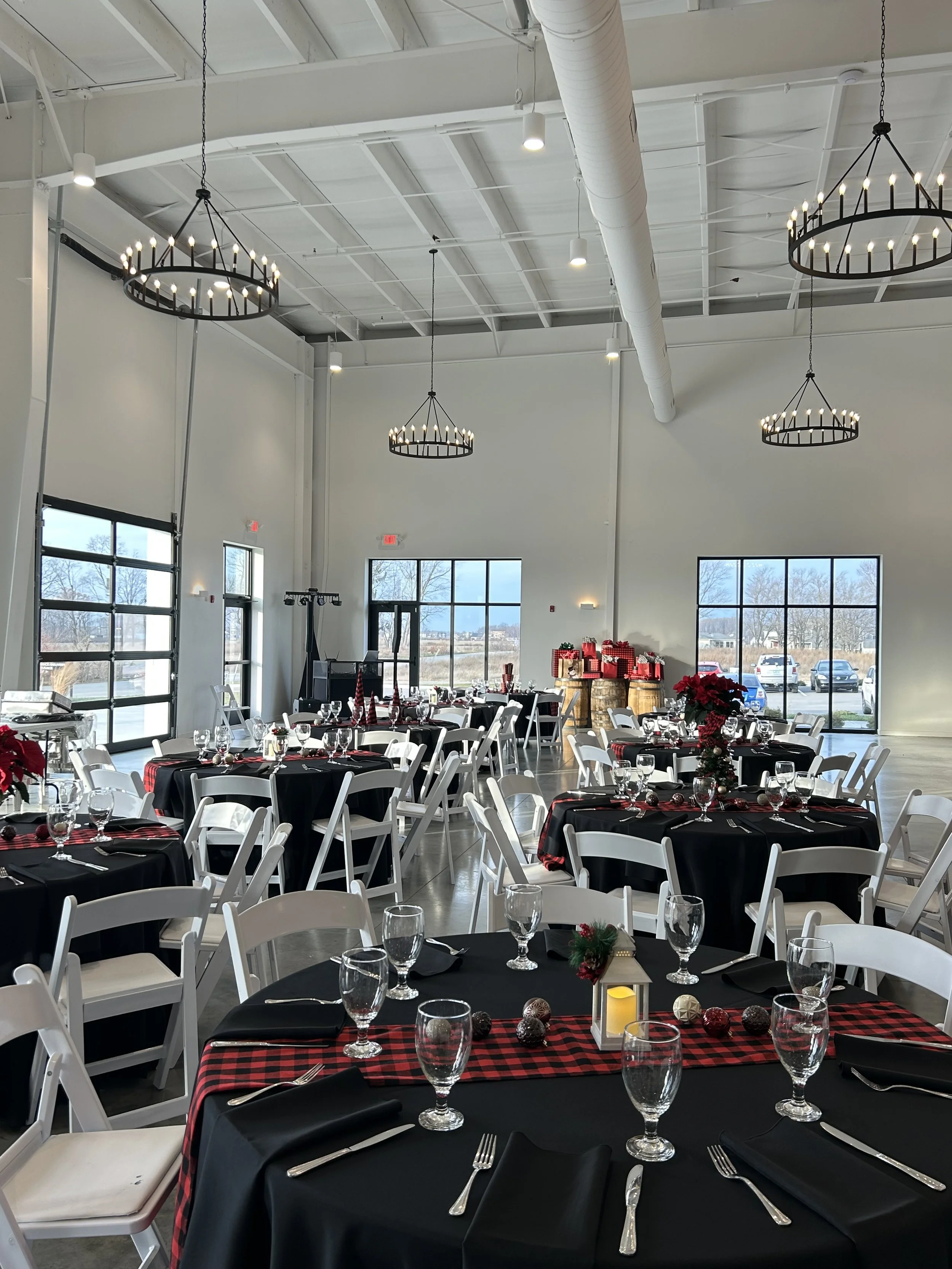 Decorated banquet hall with black and red checkered table runners, white chairs, and holiday decorations including red poinsettias, ornaments, and wrapped gifts.