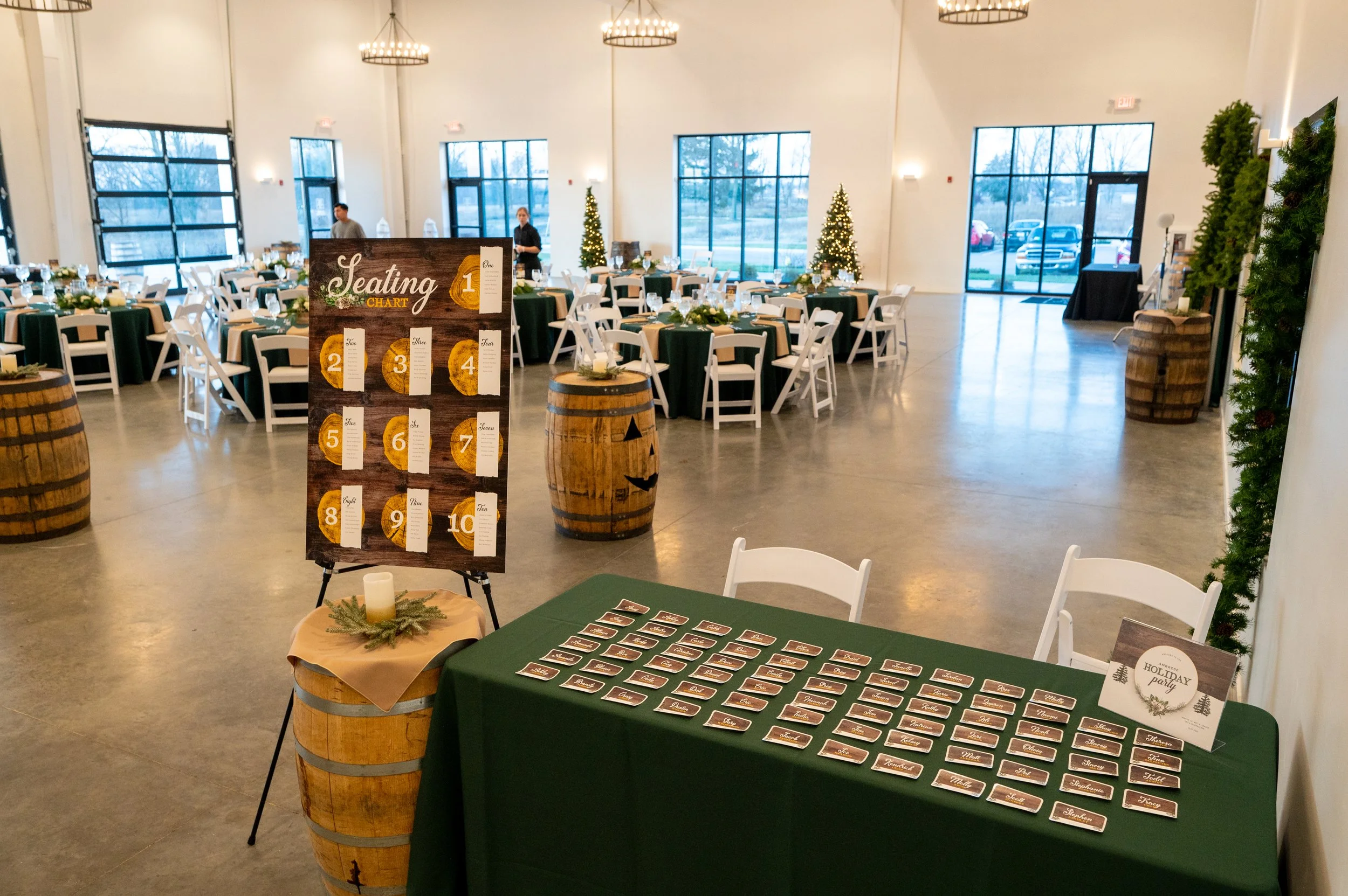 Event space decorated for a holiday gathering with trees, tables with green tablecloths, and a seating chart sign with chalkboard background featuring numbered sections on a stand and a table with name tags.