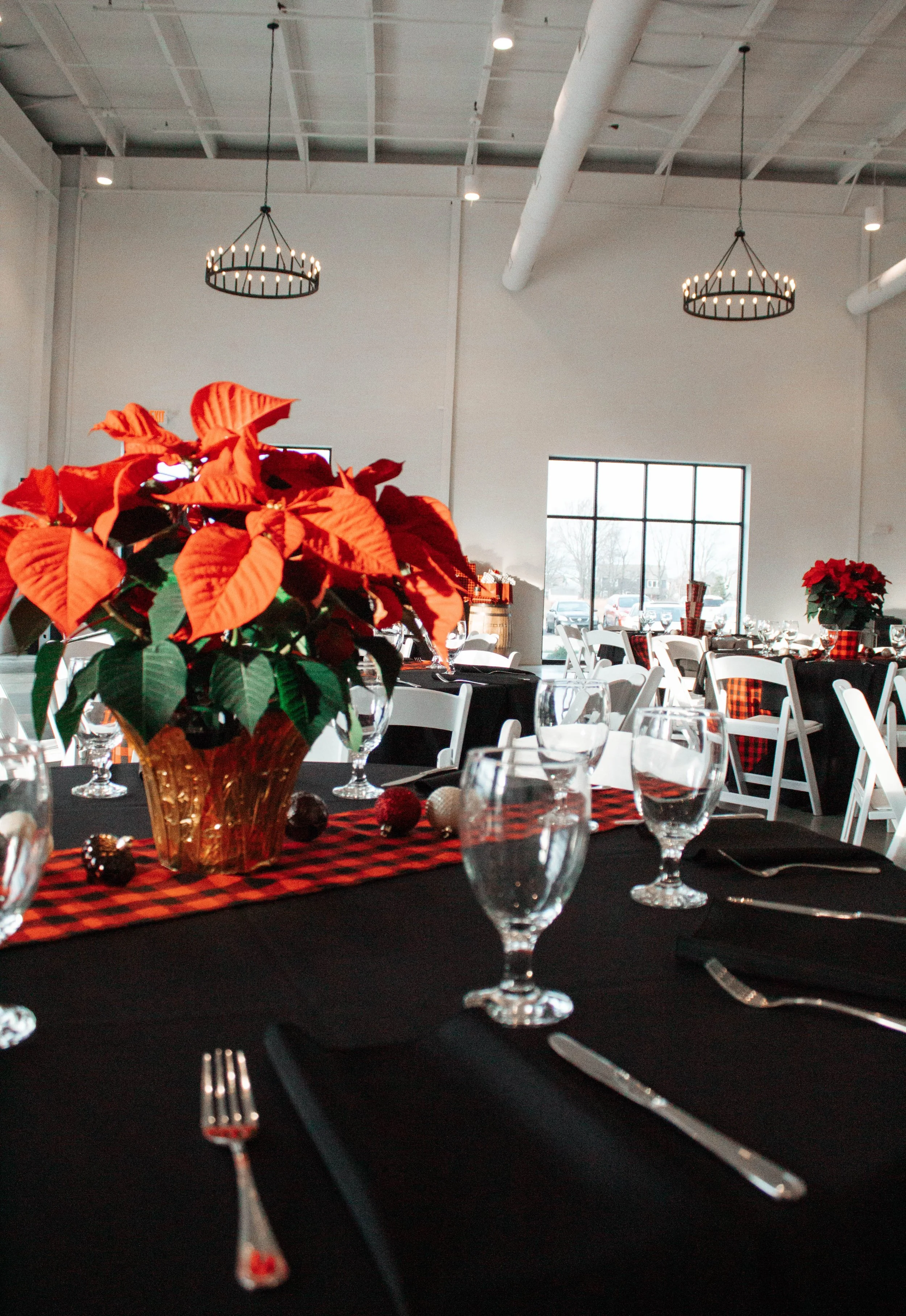 A decorated banquet hall with black tablecloths, red poinsettia flowers in gold vases, and plaid table runners, set for a festive meal.