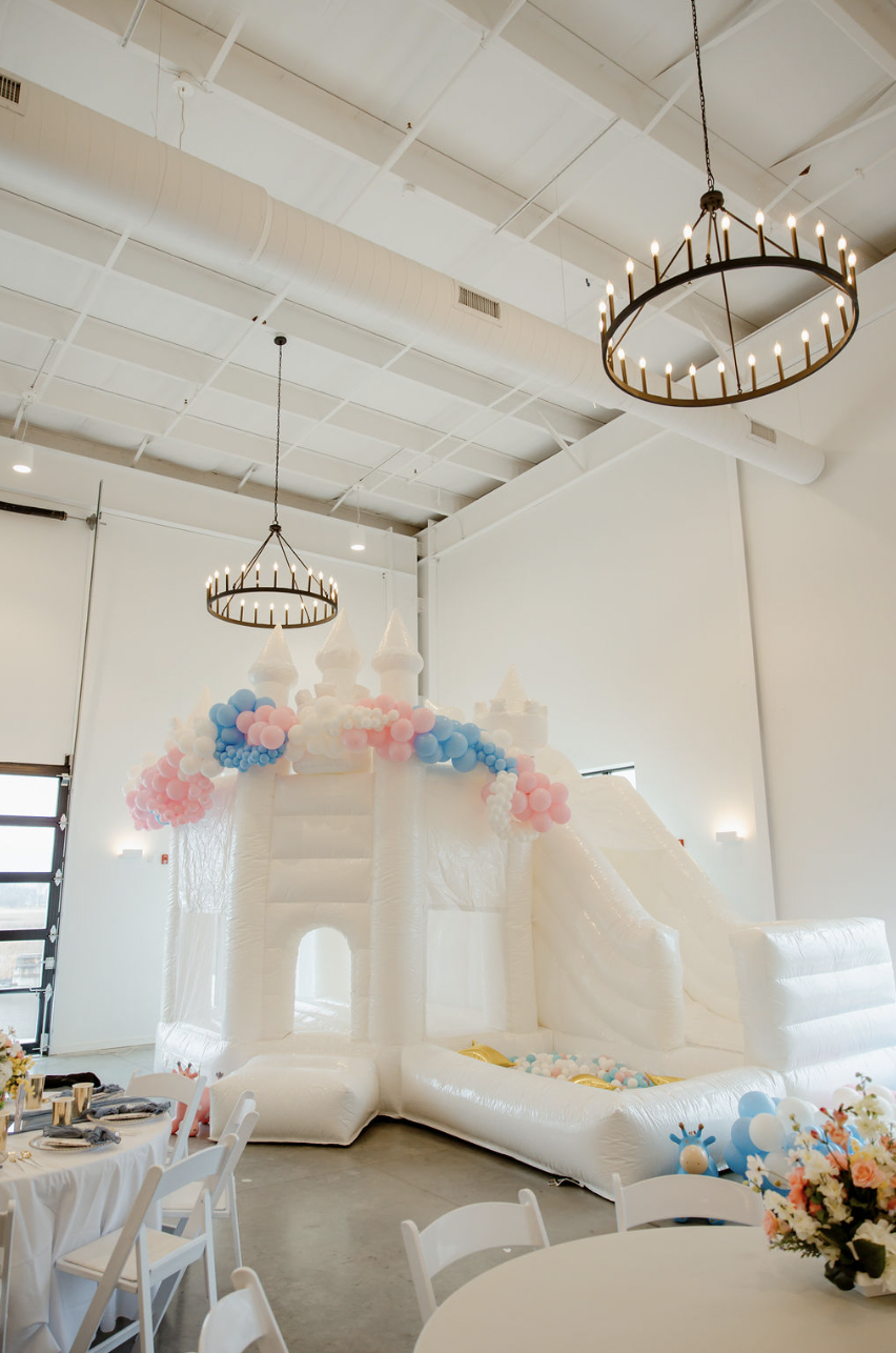 Indoor party room with white inflatable castle slide decorated with pink, blue, and white balloons, round tables with floral centerpieces, and chandeliers hanging from the ceiling.