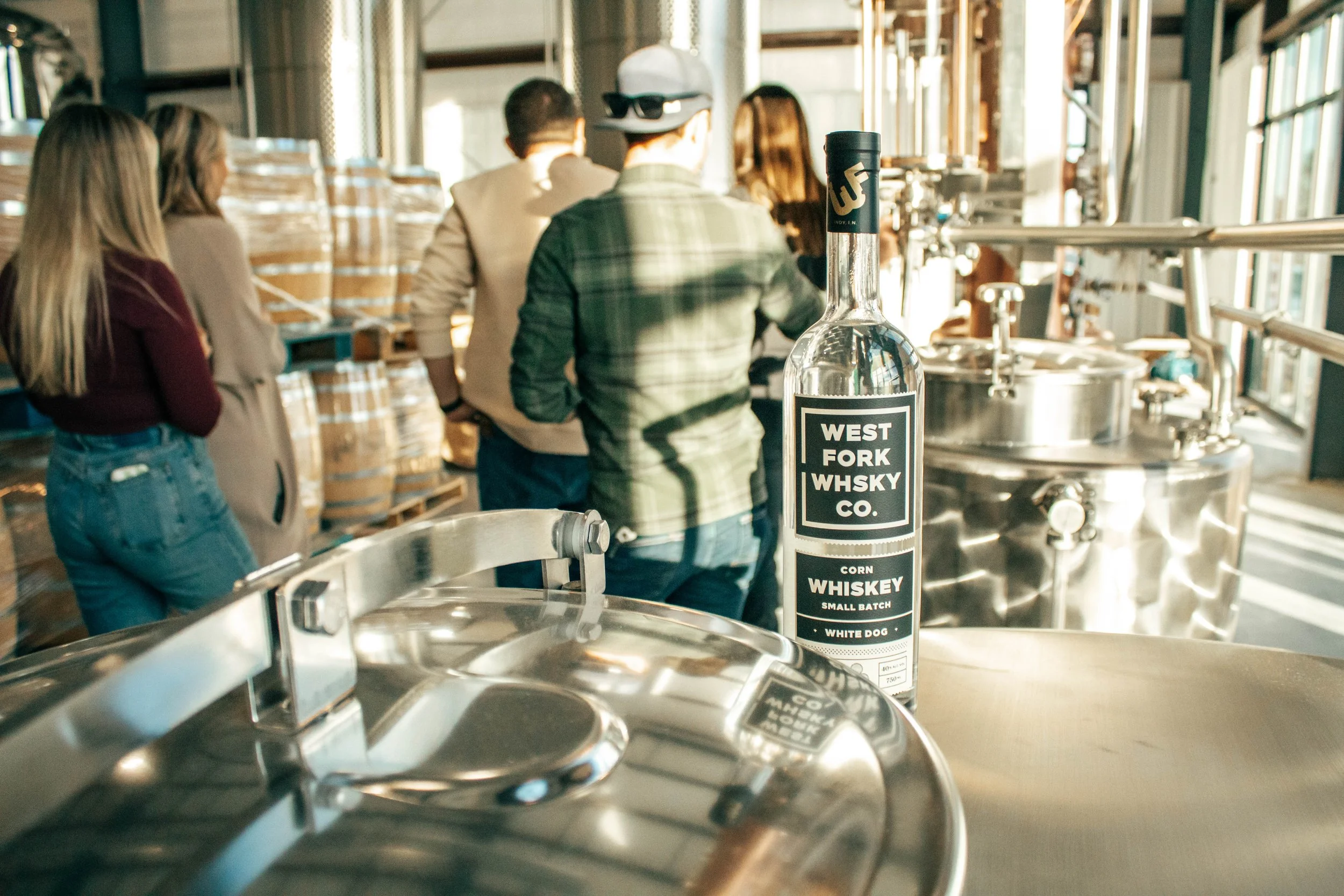 Group touring a distillery with metal equipment and whiskey bottle labeled 'West Fork Whisky Co.' in the foreground.