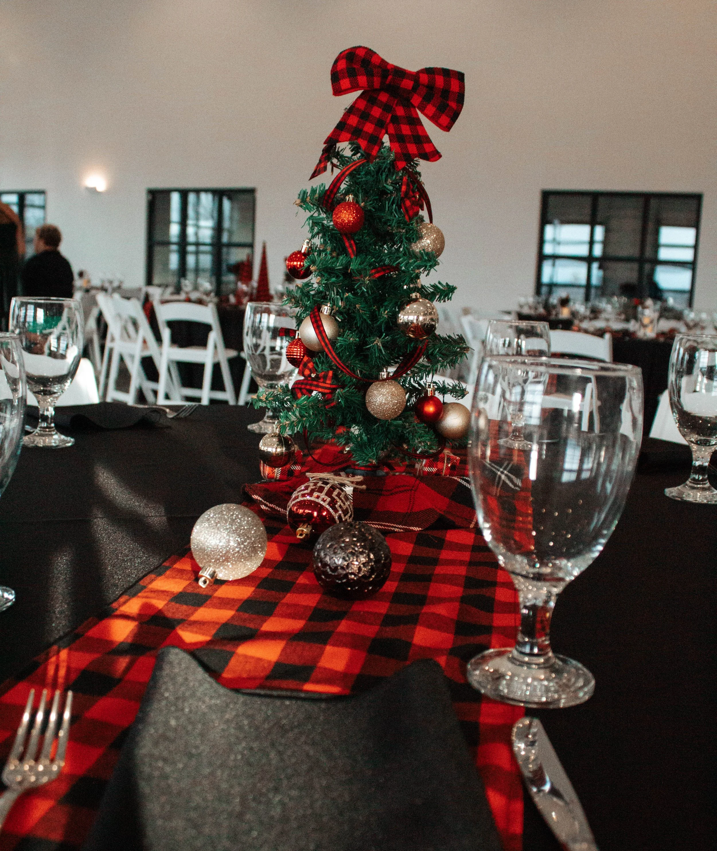 Small decorated Christmas tree with red and gold ornaments and a red and black plaid bow on top, placed on a table with a red and black plaid table runner, surrounded by empty wine glasses and black napkins.