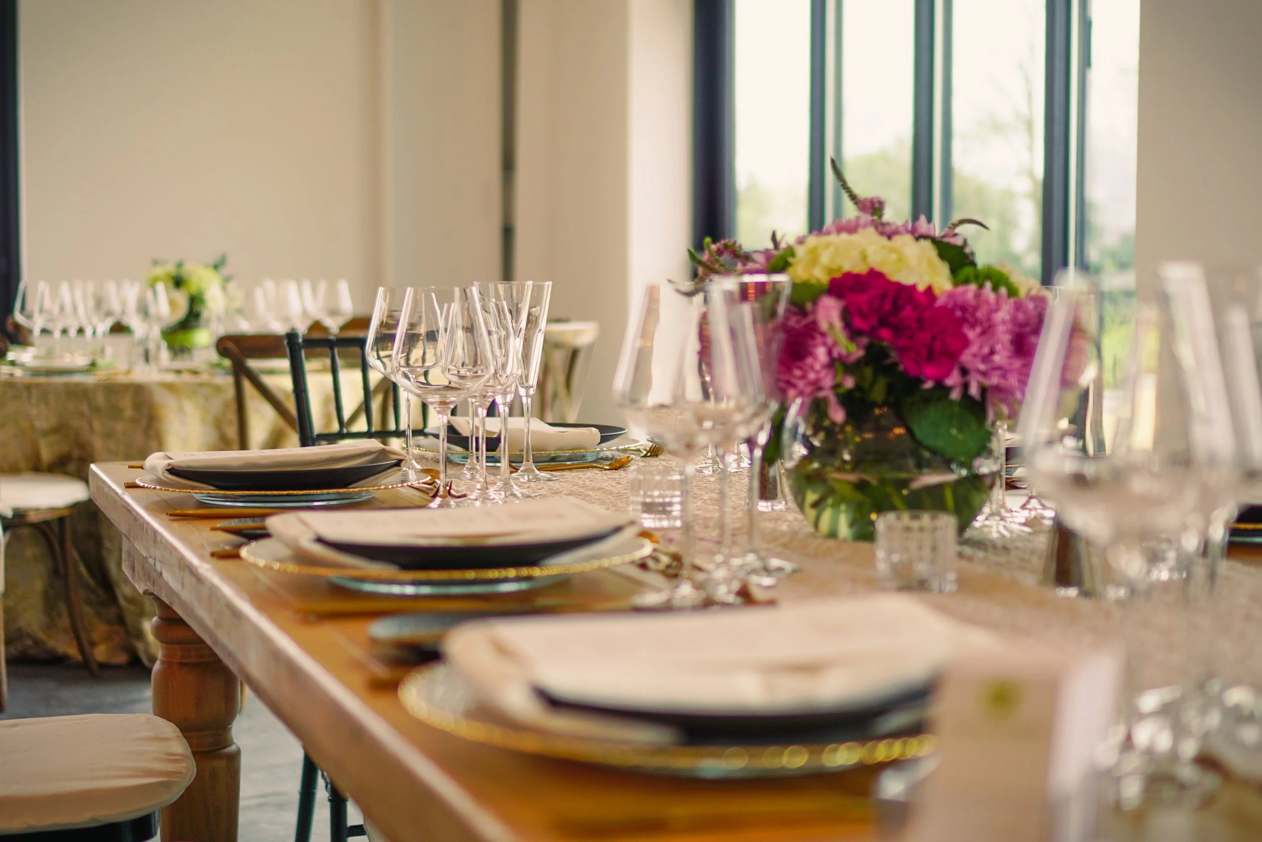Elegant dining table set with black and white plates, crystal glasses, and a floral centerpiece featuring pink, purple, and white flowers in a glass vase, near large windows.